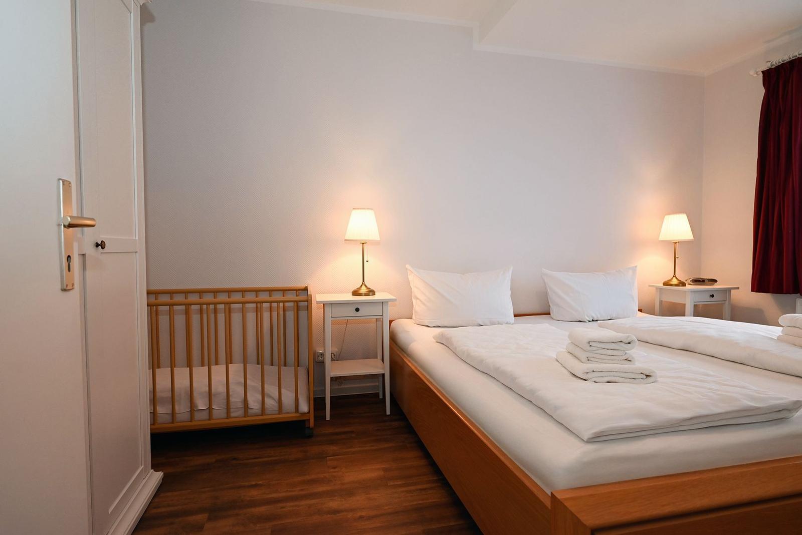 Schlafzimmer mit Doppelbett, Kinderbett, zwei Nachttischen und rotem Vorhang.