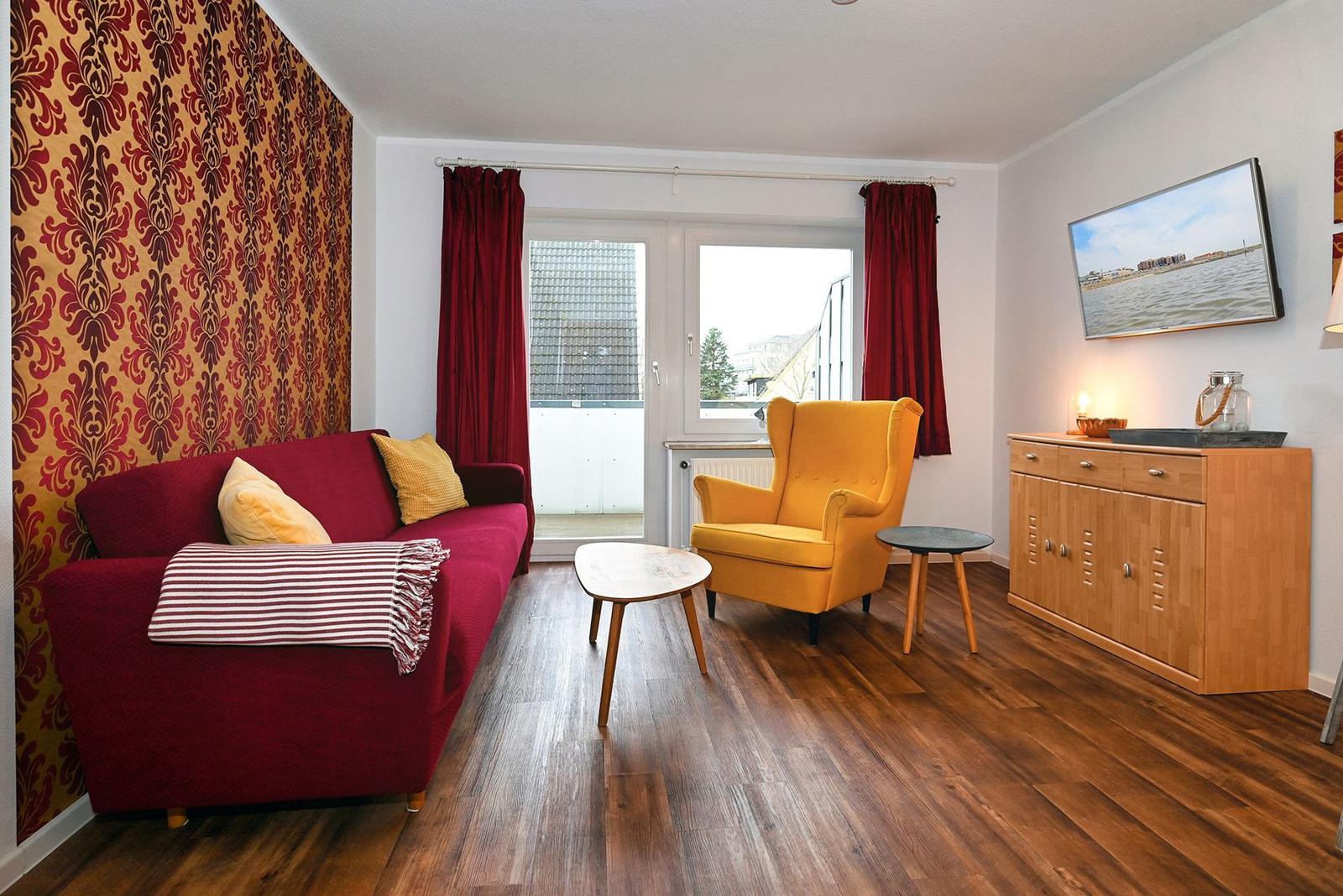 Ein Wohnzimmer mit rotem Sofa, gelbem Sessel und Holzboden. An der Wand hängt ein Fernseher.
