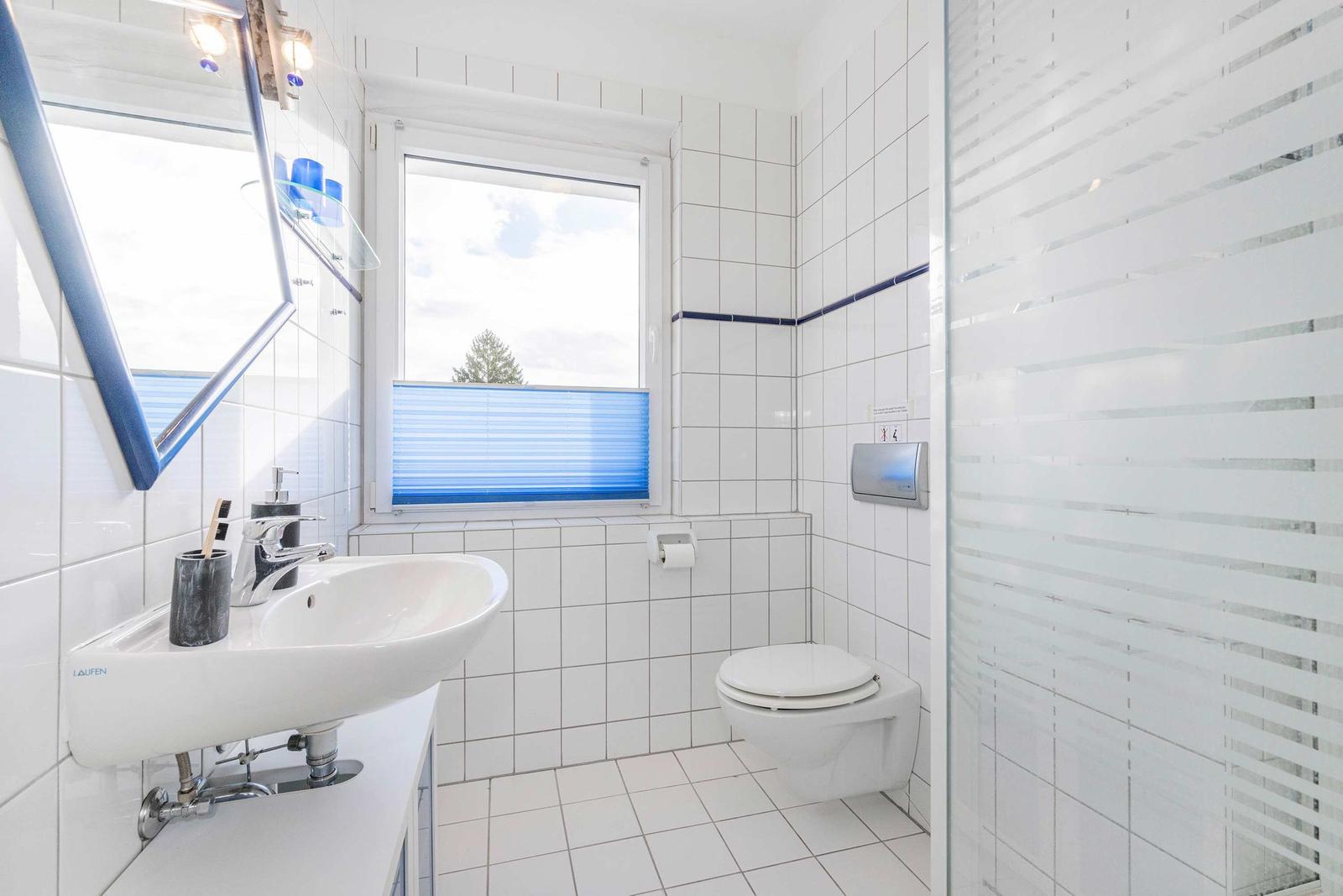 Badezimmer mit Waschbecken, Toilette und Dusche. Fenster mit blauen Jalousien.