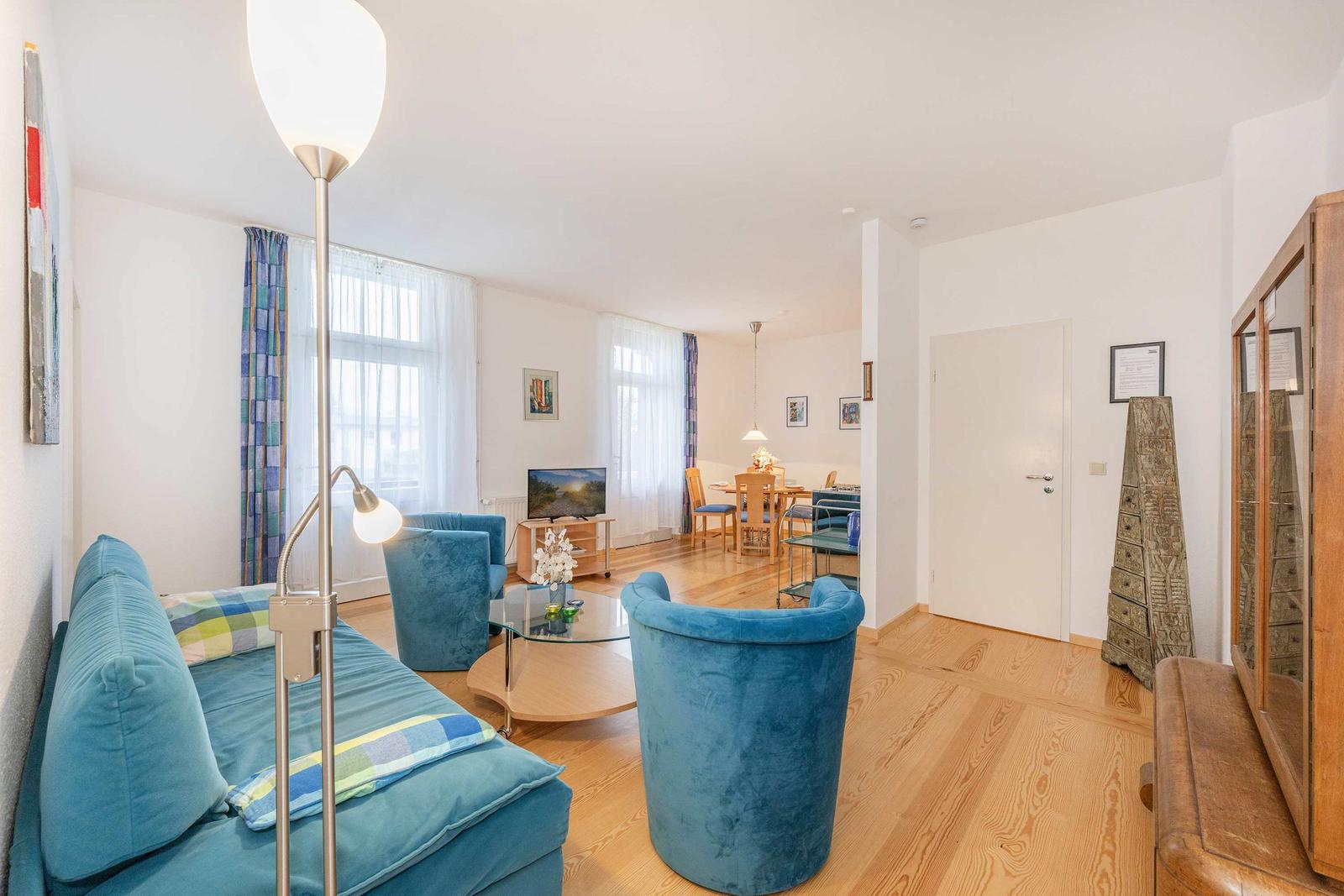 Wohnzimmer mit blauer Couch und Sesseln, Glas-Tisch und Holzboden. Essbereich im Hintergrund.