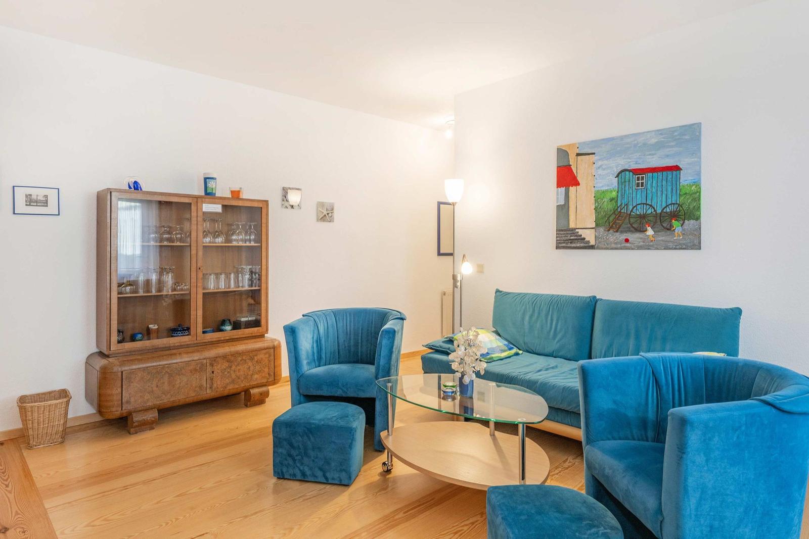 Wohnzimmer mit blauen Sesseln, Sofa und Glas-Tisch. Holzschrank und Wandbild sichtbar.