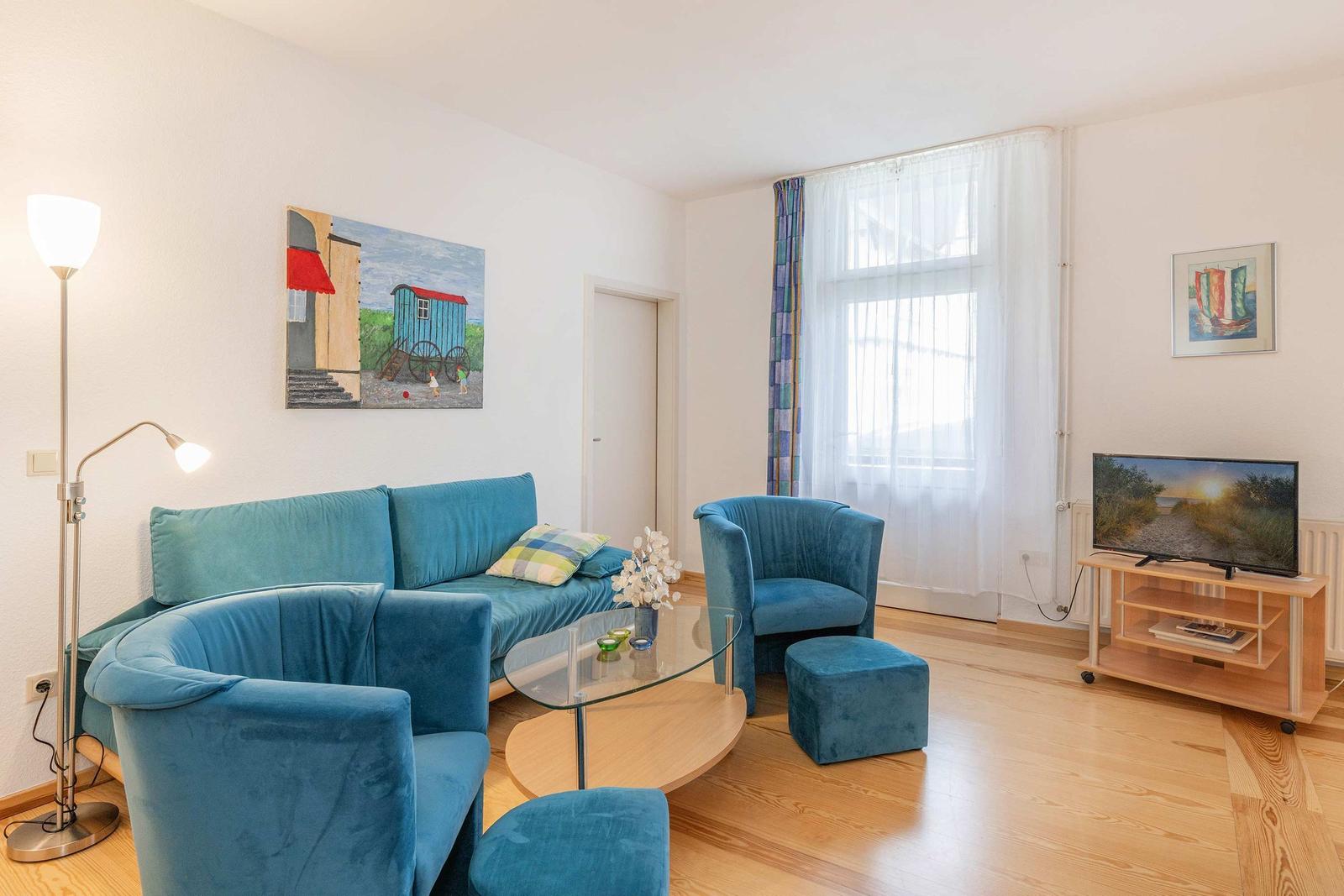 Wohnzimmer mit blauer Couch, Sessel und Glas-Tisch. Fenster mit weißen Vorhängen.