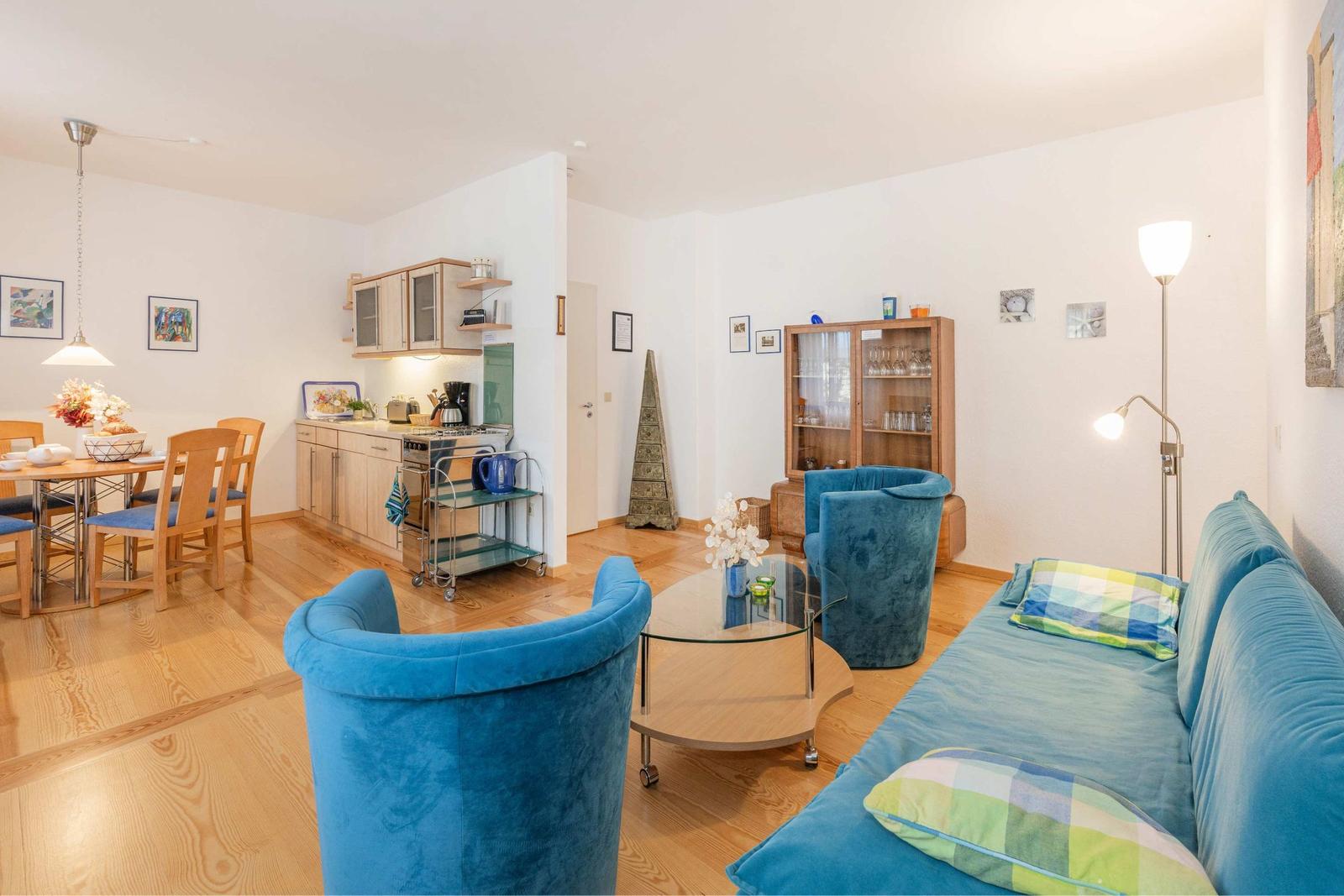 Wohnbereich mit blauer Couch, zwei Sesseln und Glas-Tisch. Küche und Essbereich sind sichtbar.