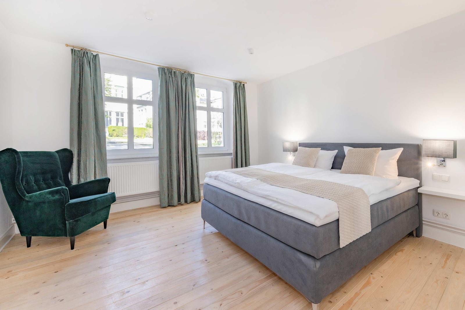Schlafzimmer mit Bett, grüner Sessel und Fenster mit Vorhängen.