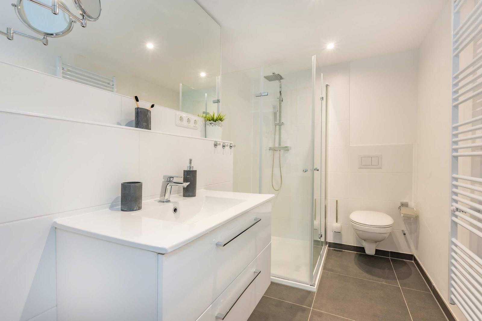 Badezimmer mit Dusche, Waschbecken und Toilette. Weiße Wandfliesen und graue Bodenfliesen.