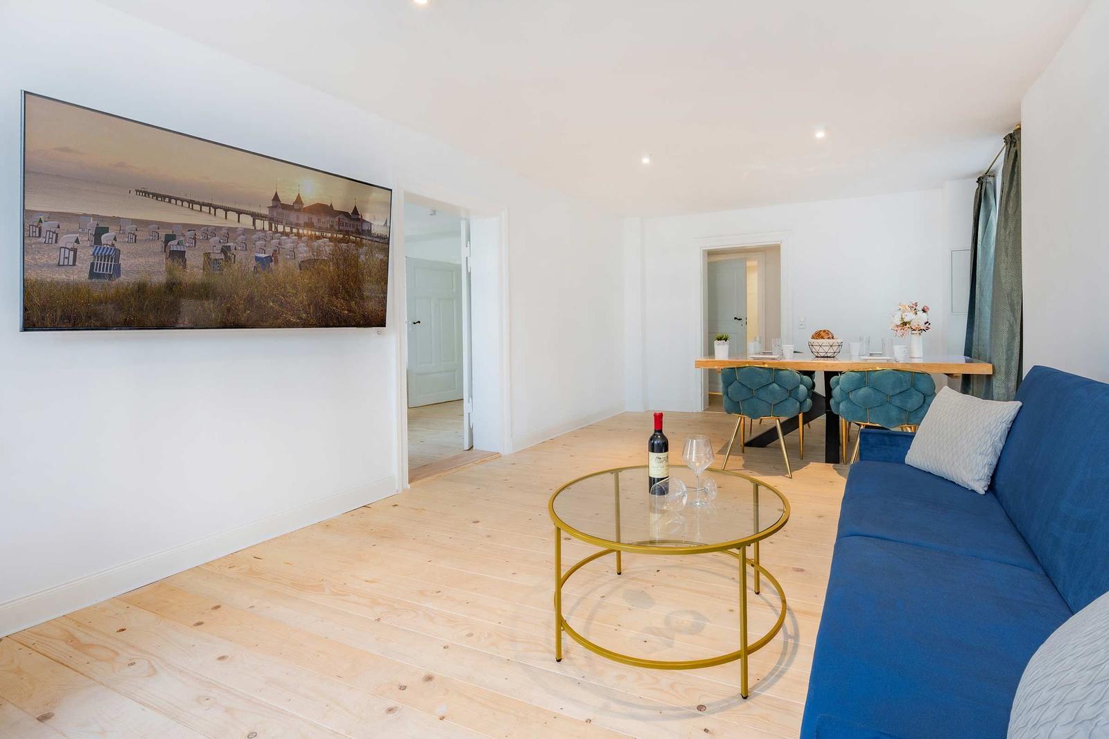 Wohnzimmer mit blauem Sofa, Glas-Tisch und Wandbild. Essbereich mit blauen Stühlen.