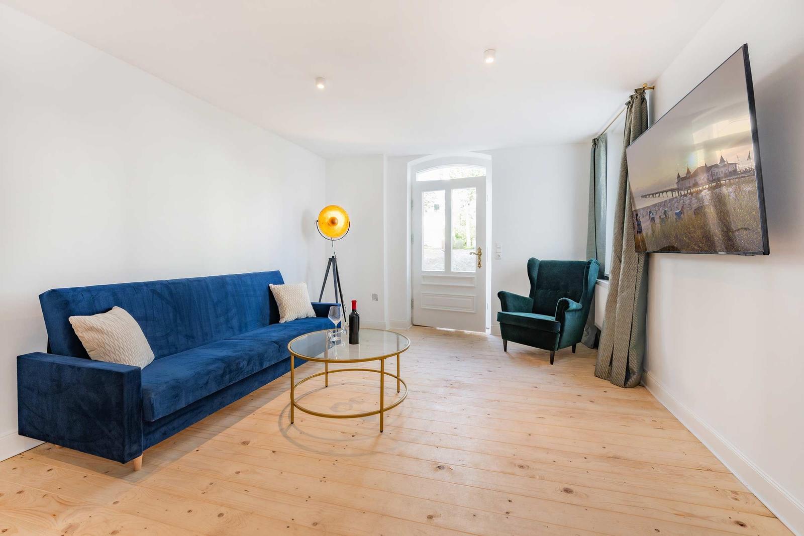 Wohnzimmer mit blauem Sofa, grünem Sessel und Glas-Tisch auf Holzboden.