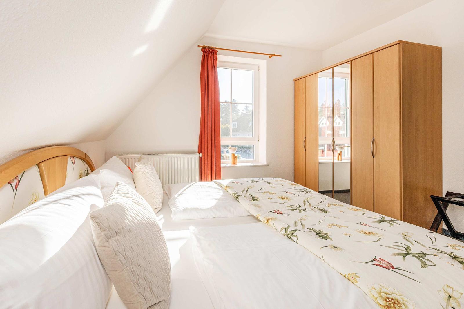 Schlafzimmer mit Dachschrägen, Bett, Kleiderschrank und Fenster mit roter Gardine.