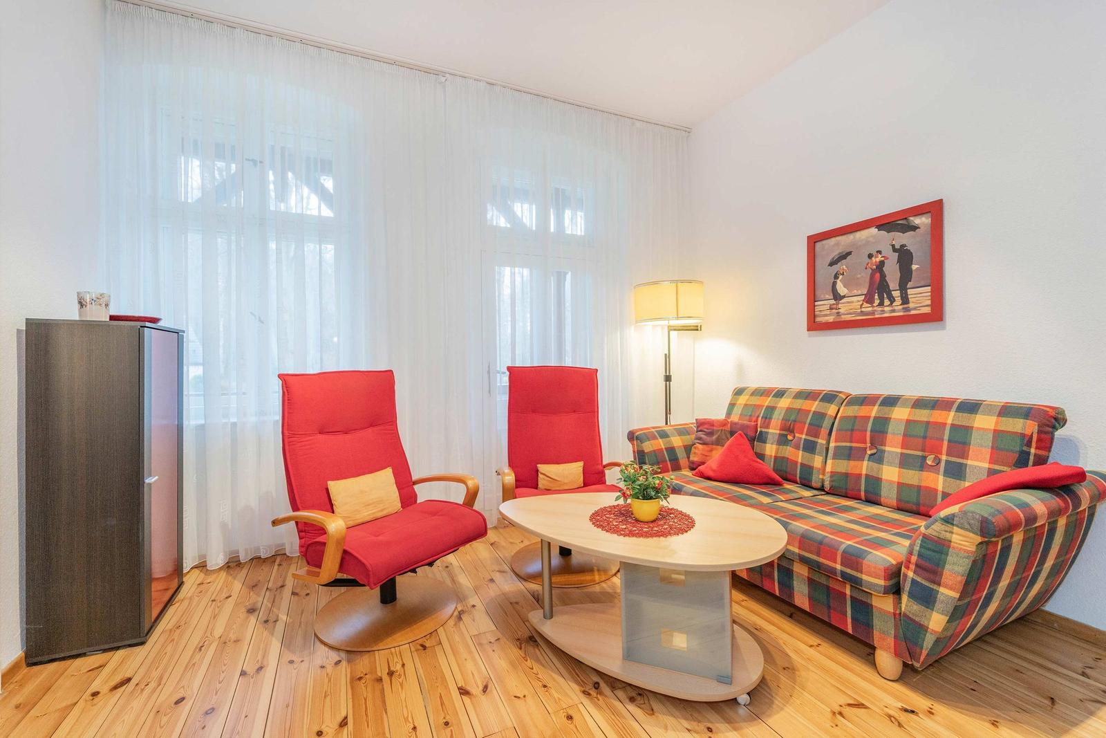 Wohnzimmer mit Sofa, zwei roten Sesseln, Tisch und Fenster mit Vorhängen.