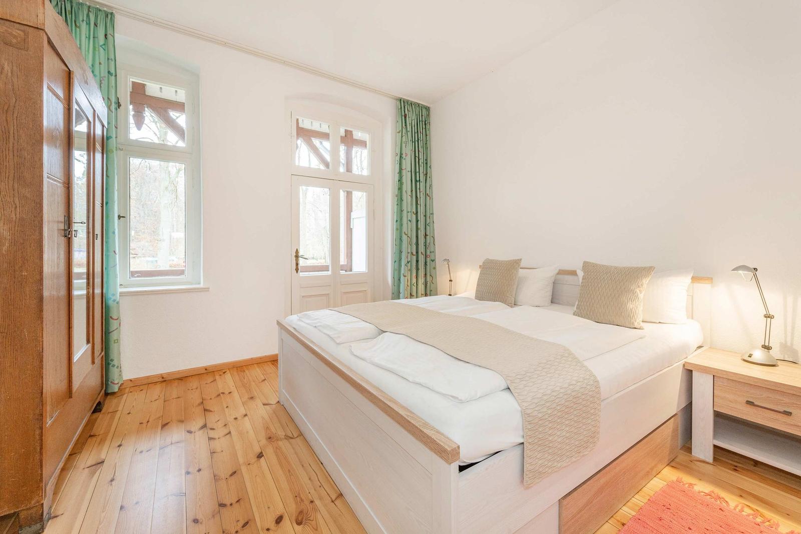 Schlafzimmer mit Doppelbett, Holzboden und Fenstern mit grünen Vorhängen.
