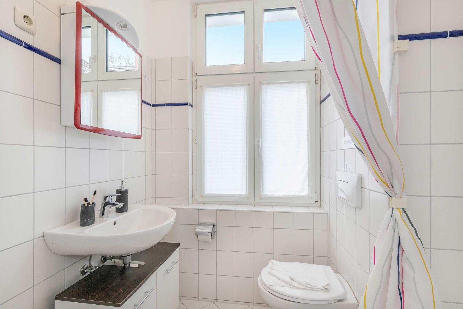 Badezimmer mit Waschbecken, Spiegel, Toilette und Fenster mit Jalousie.