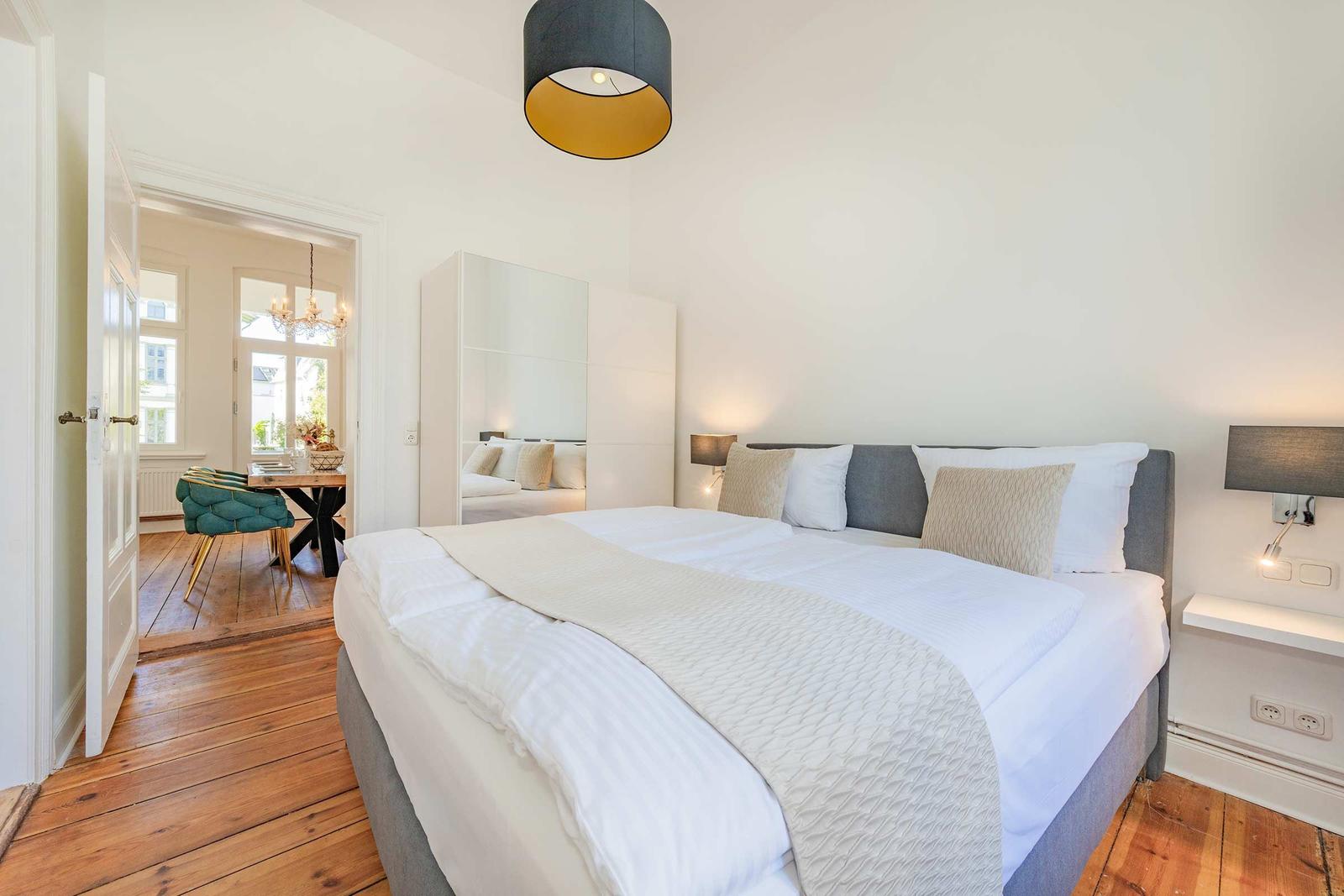 Schlafzimmer mit Doppelbett, Spiegelschrank und Holzboden. Durchgang zu Essbereich mit Tisch und Stuhl.