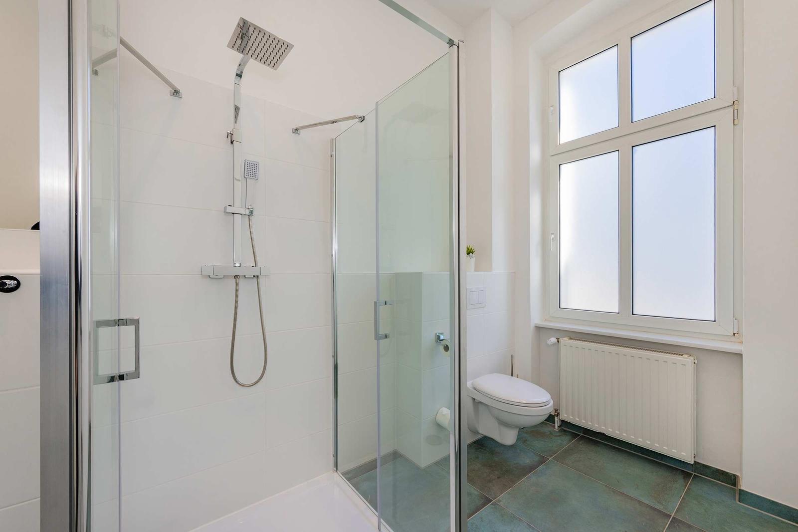 Duschkabine mit Duschbrause, Toilette und Fenster mit Heizkörper darunter.