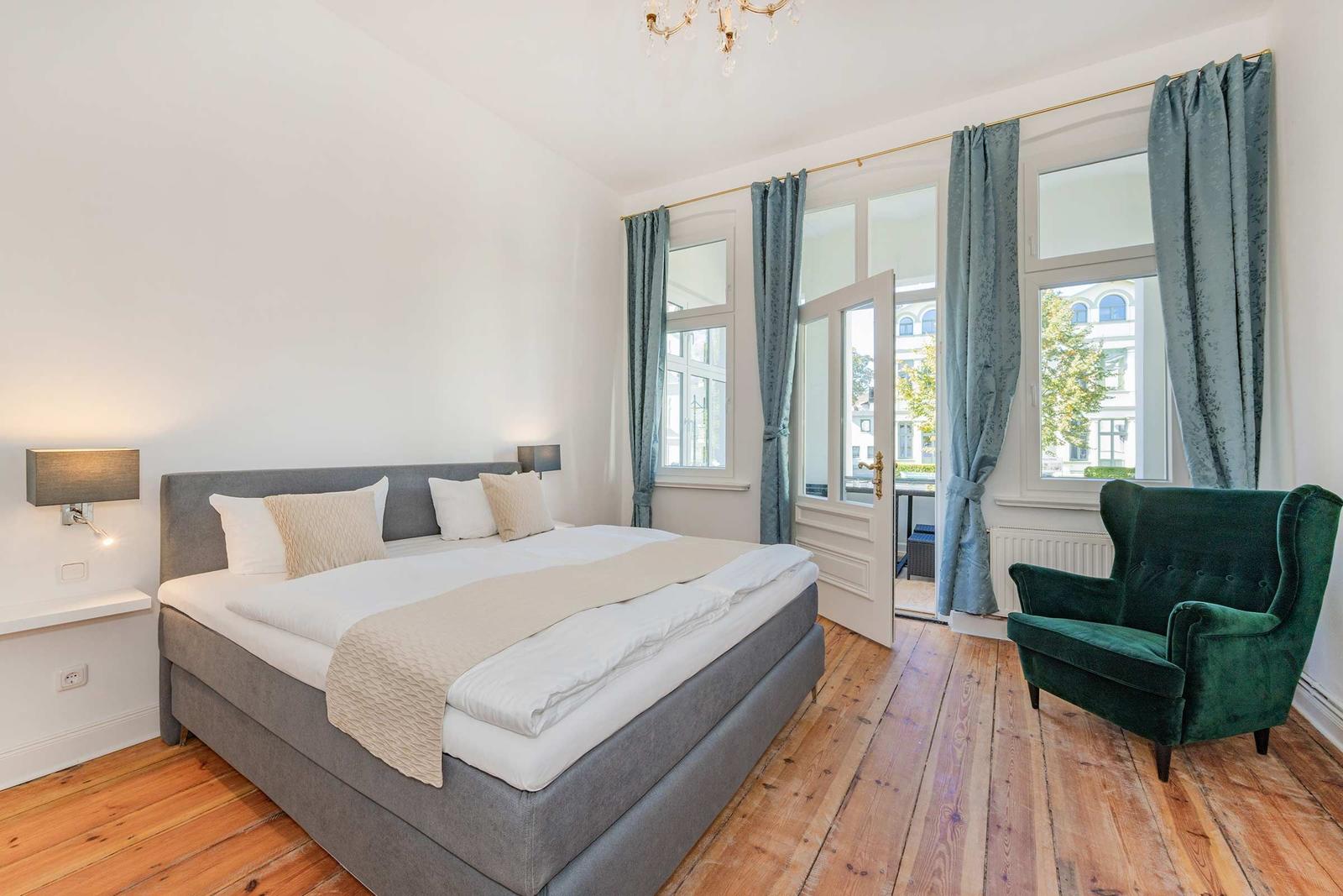 Schlafzimmer mit Bett, grüner Sessel und Holzboden. Fenster mit Vorhängen und Tür zum Balkon.