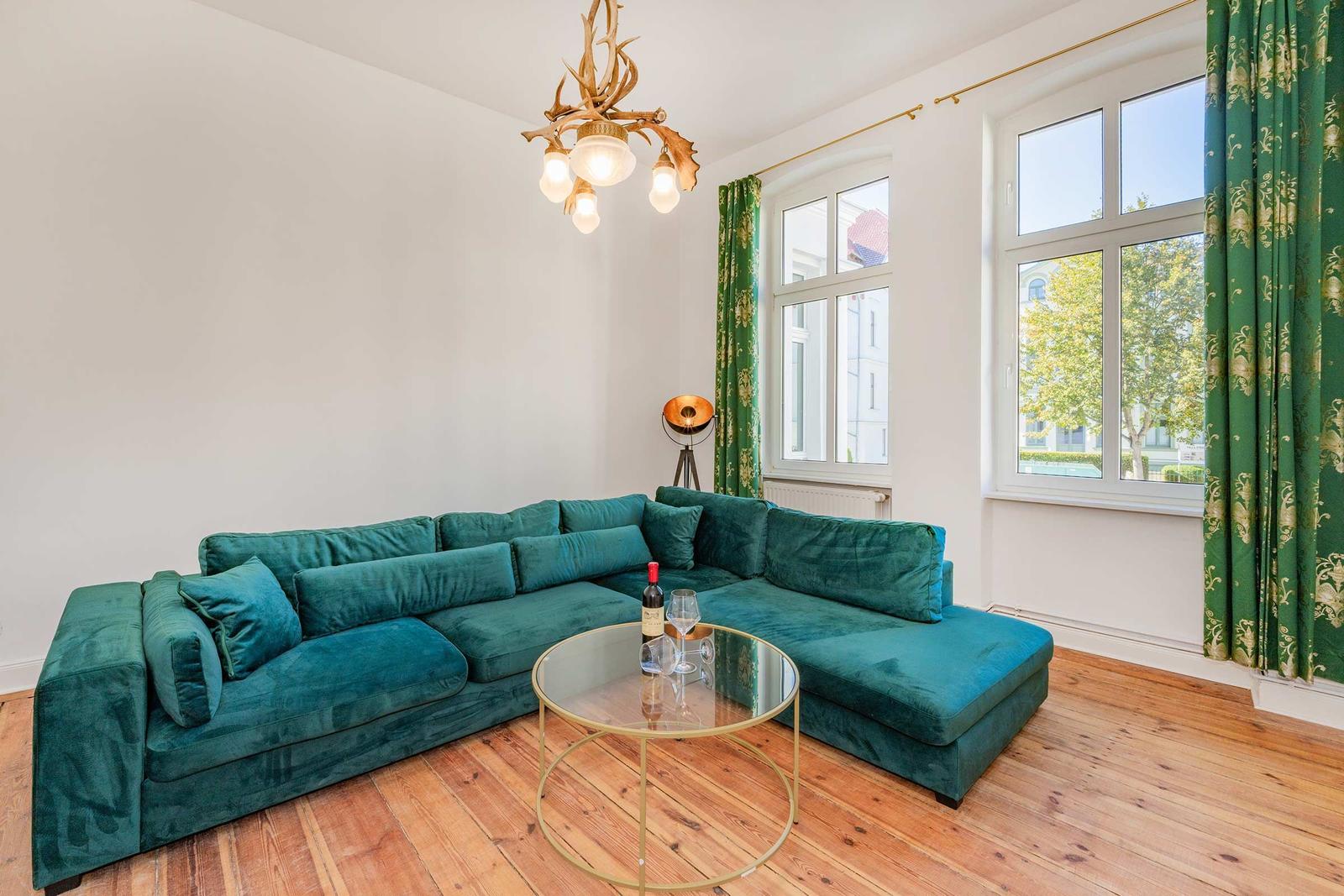Ein Wohnzimmer mit grünem Sofa, Holzboden und großen Fenstern mit grünen Vorhängen.