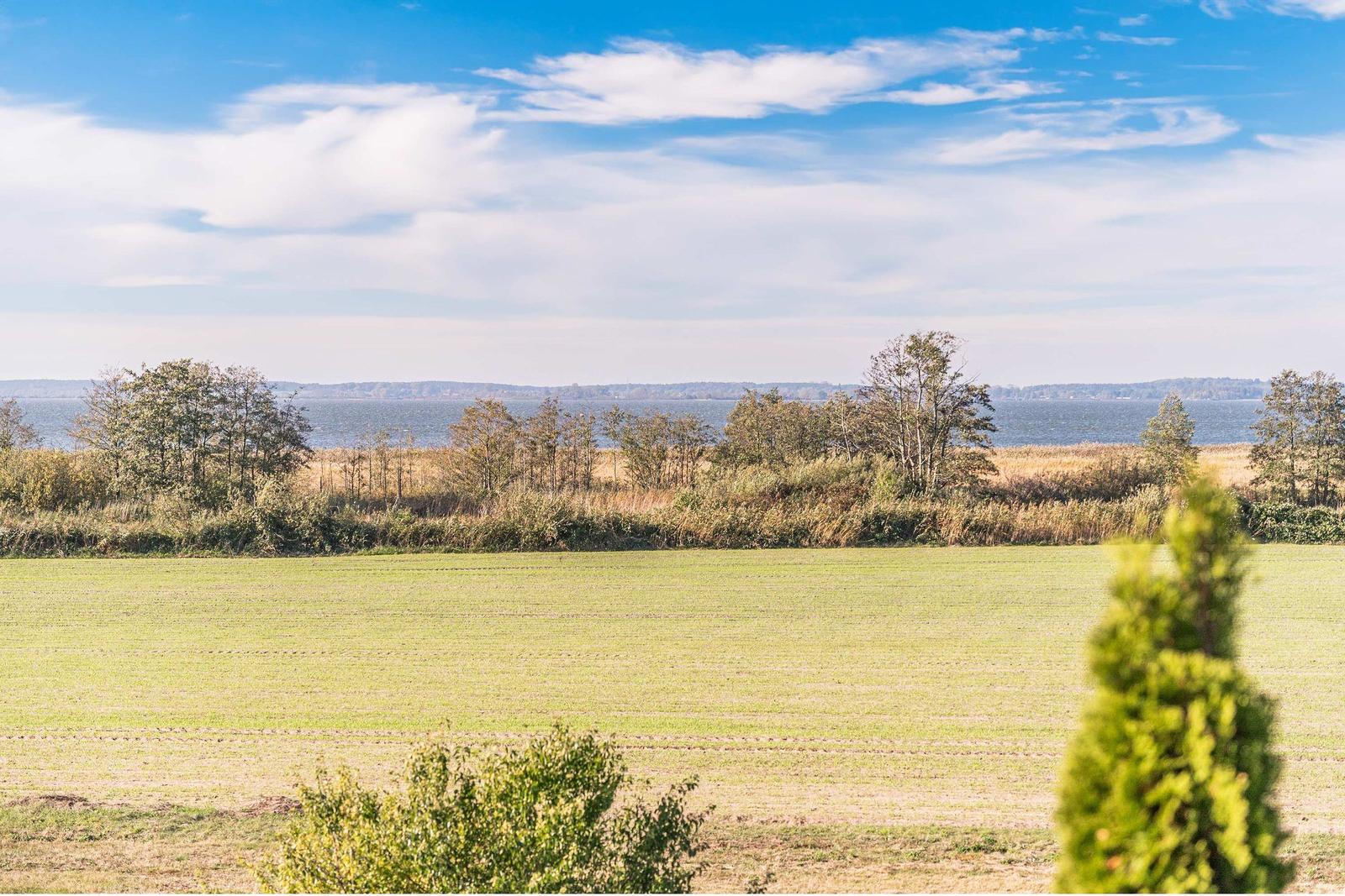 Weitläufiges Feld mit Blick auf einen See und Bäume im Hintergrund unter blauem Himmel.