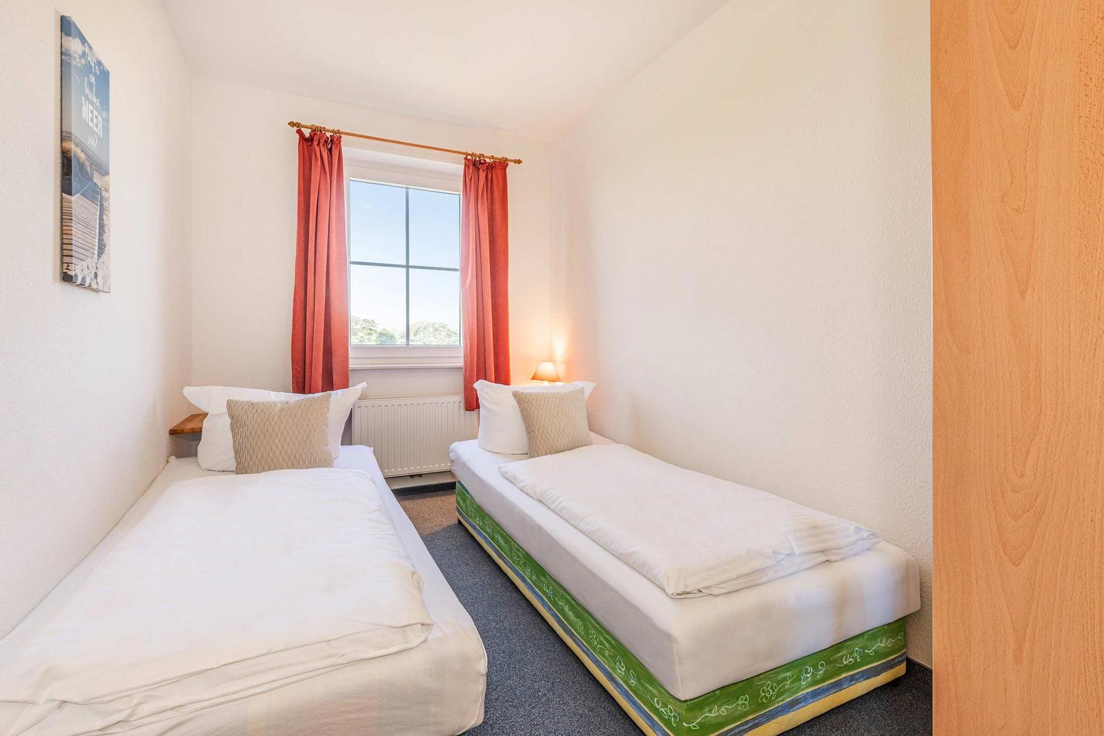 Zwei Einzelbetten mit weißen Bettwäschen und Kissen. Fenster mit roten Vorhängen und Wandbild.