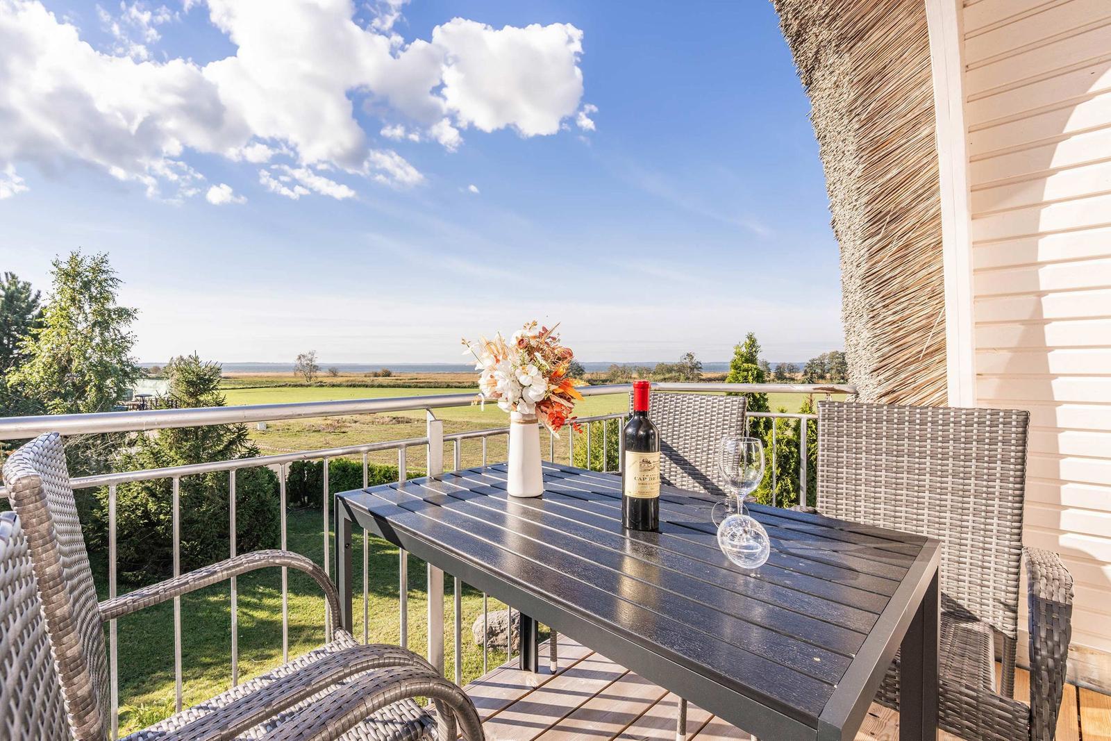 Terrasse mit Tisch, Stühlen, Weinflasche und Blumen. Blick auf Felder und Himmel.