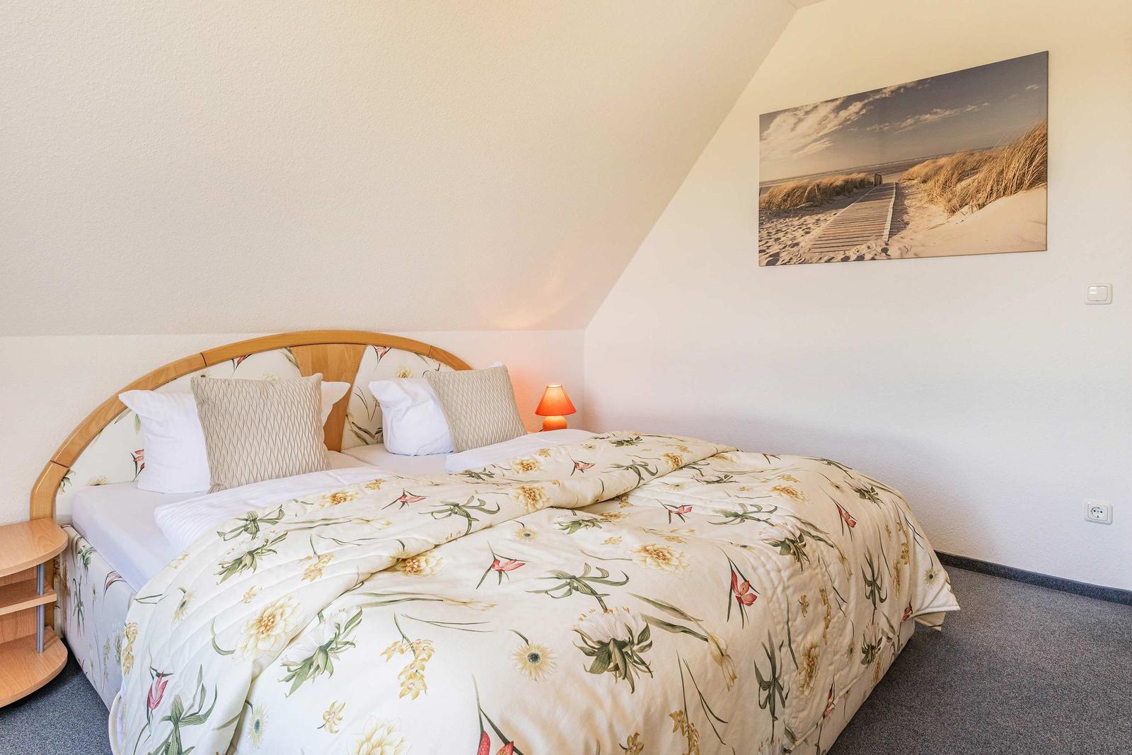 Schlafzimmer mit Doppelbett, floraler Bettwäsche und Wandbild an der Schrägwand.