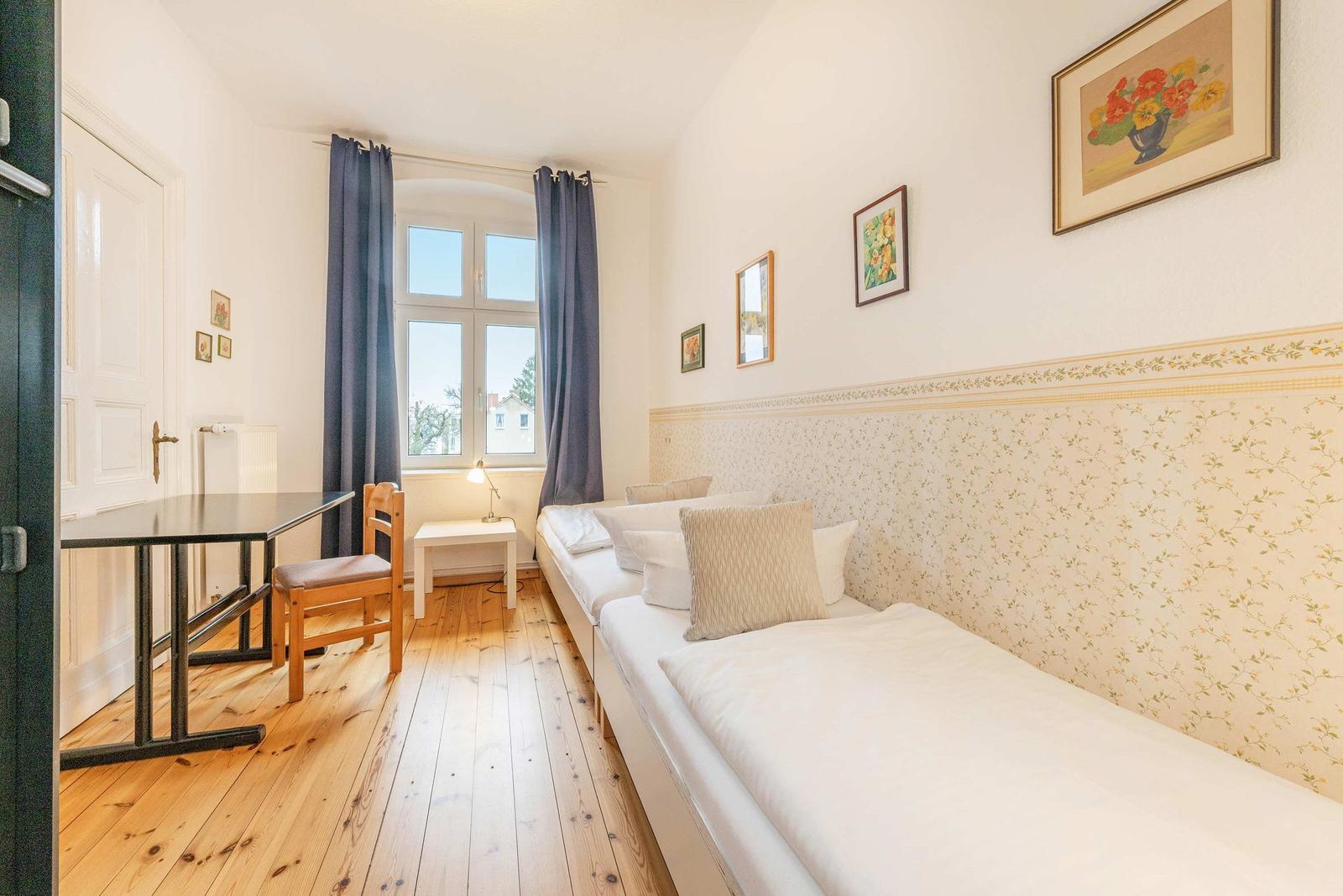 Zimmer mit zwei Betten, Holzboden, Tisch, Stuhl und Fenster mit Vorhängen.