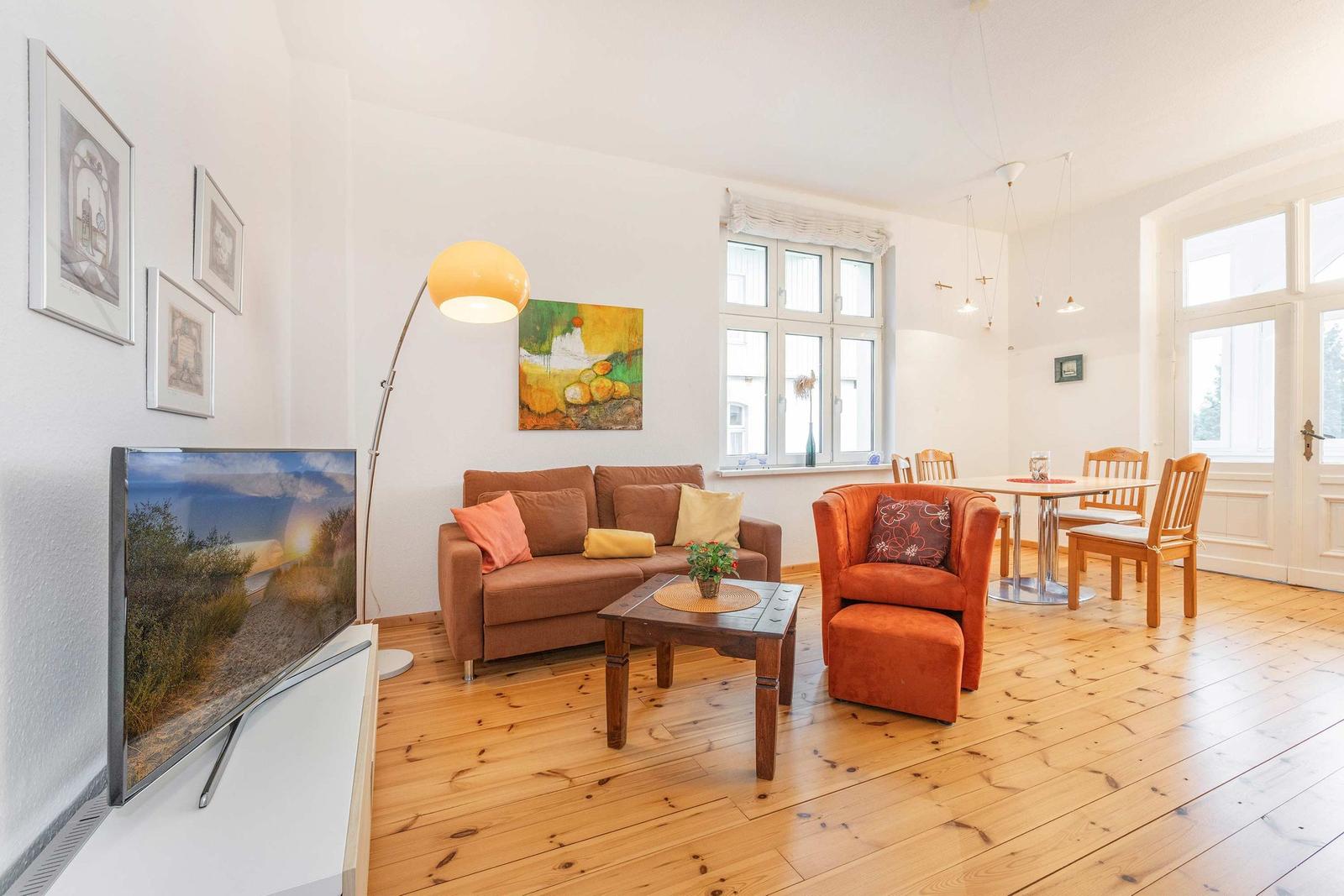 Wohn- und Essbereich mit Sofa, Sessel, Tisch und TV. Holzfußboden und große Fenster.