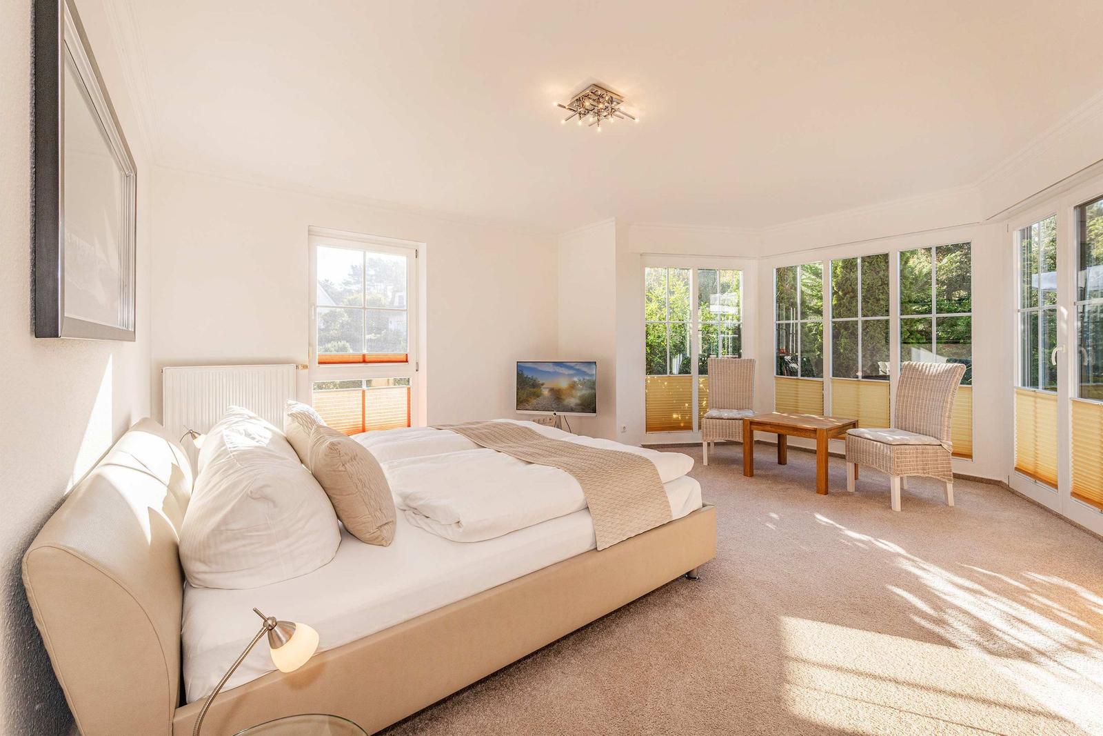 Schlafzimmer mit Doppelbett, TV und Sitzgelegenheit. Große Fenster mit Blick auf Garten.