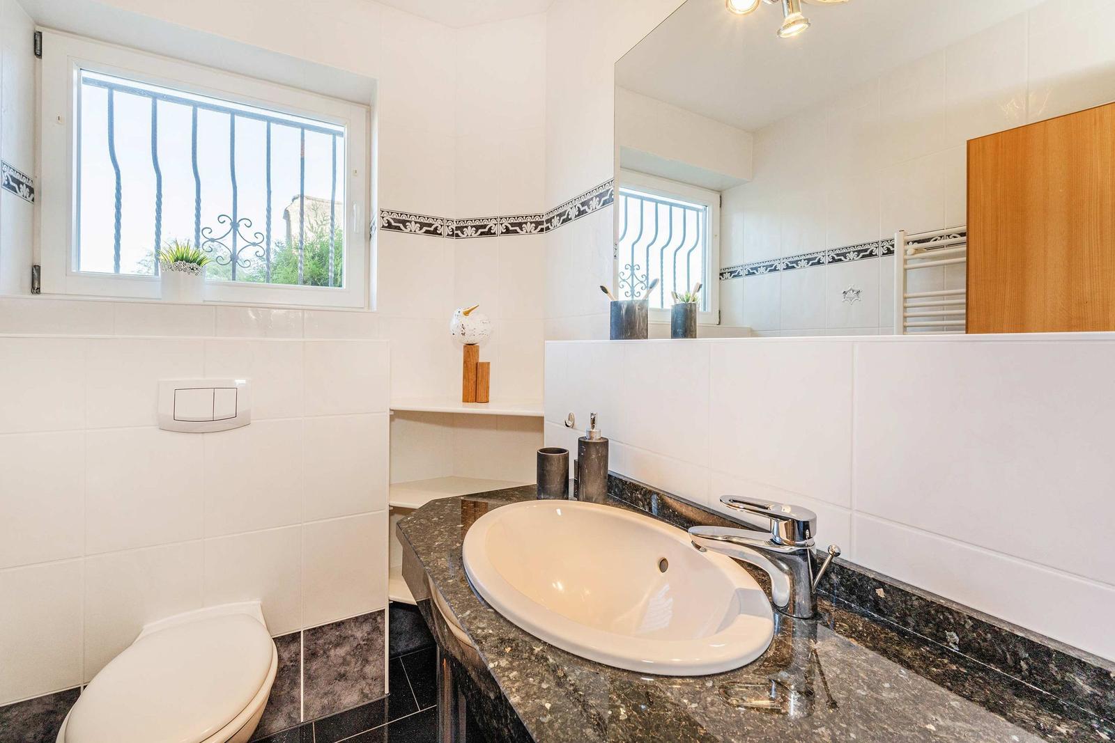 Badezimmer mit Waschbecken, Spiegel und Toilette. Fenster mit Gitter und Mosaikfliesen.