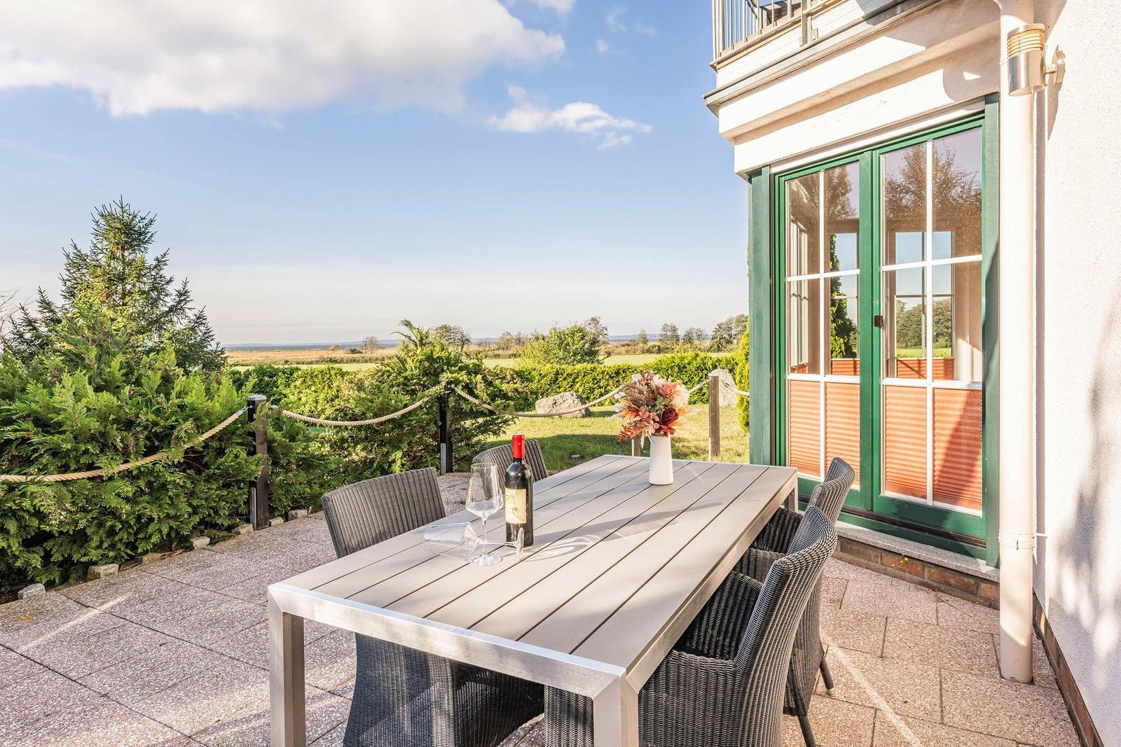 Terrasse mit Tisch und Stühlen, Blick auf grüne Landschaft und Haus mit grünen Fensterrahmen.