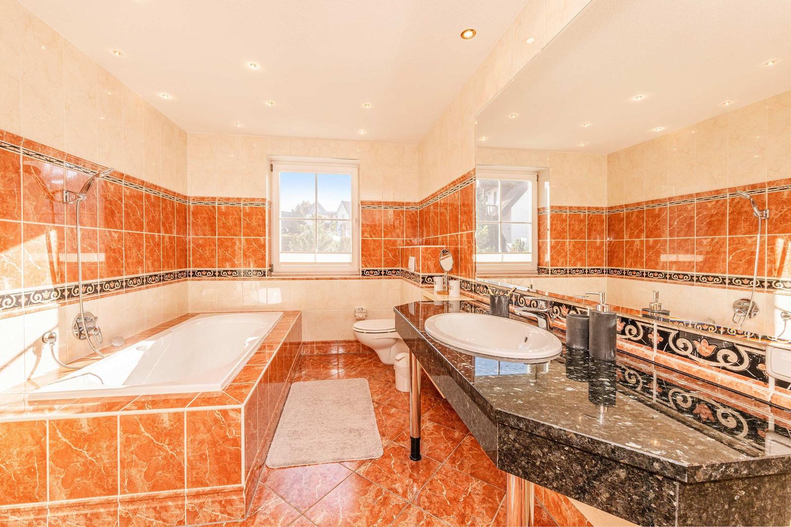 Badezimmer mit Badewanne, Waschtisch und Toilette. Wand- und Bodenfliesen in warmen Tönen.