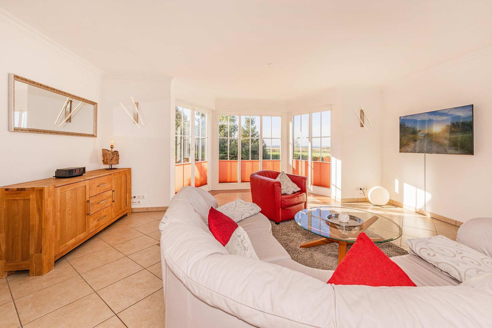 Wohnzimmer mit Sofa, rotem Sessel und Glas-Tisch. Fenster mit Blick auf Landschaft.
