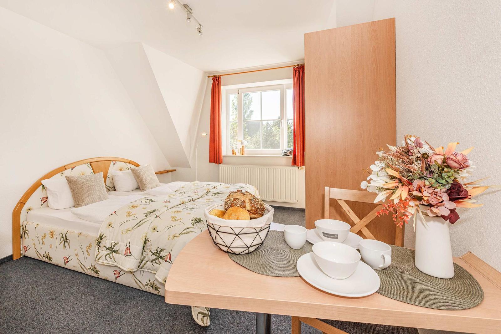 Schlafzimmer mit Dachschräge, Bett, Fenster und Esstisch mit Tasse und Blumen.