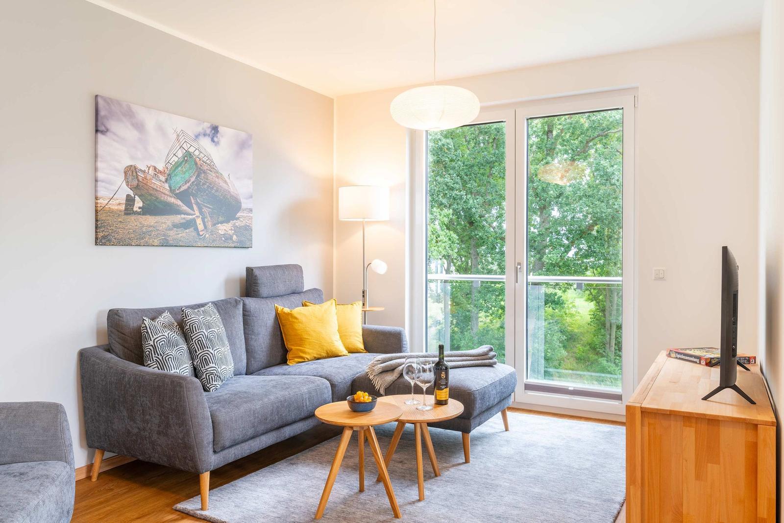 Ein Wohnzimmer mit grauem Sofa, Holztisch und Balkon mit Blick auf Bäume.