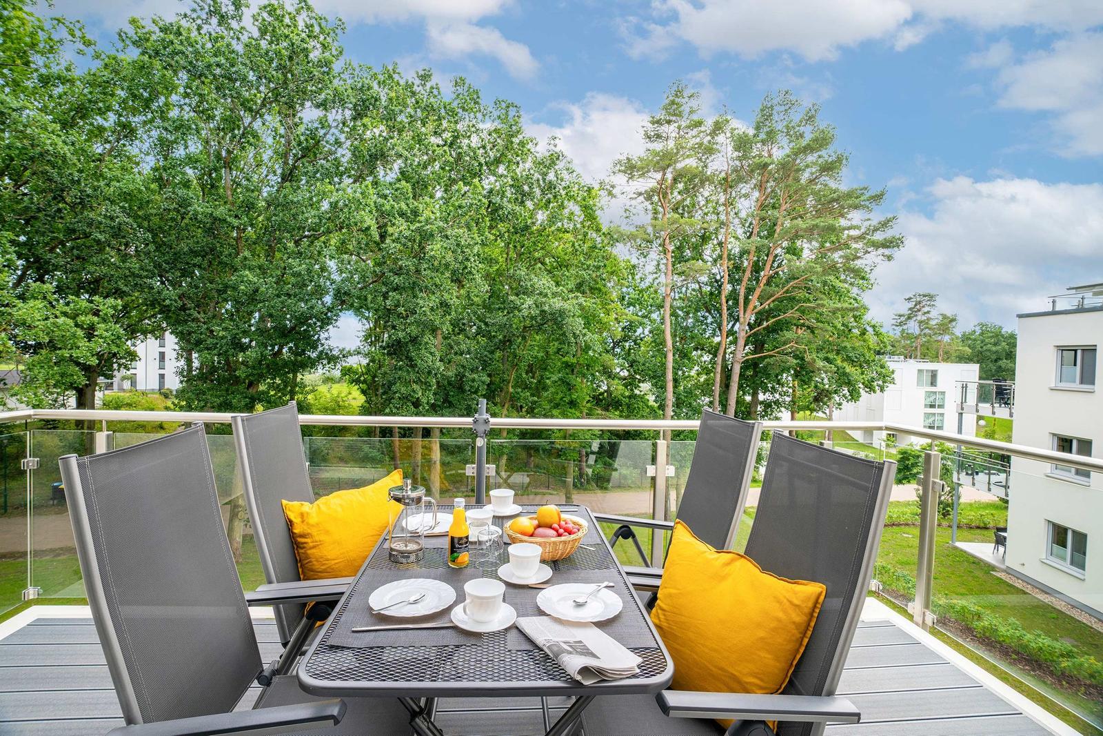 Terrasse mit Tisch, Stühlen und gelben Kissen. Blick auf Bäume und Wohnhäuser.