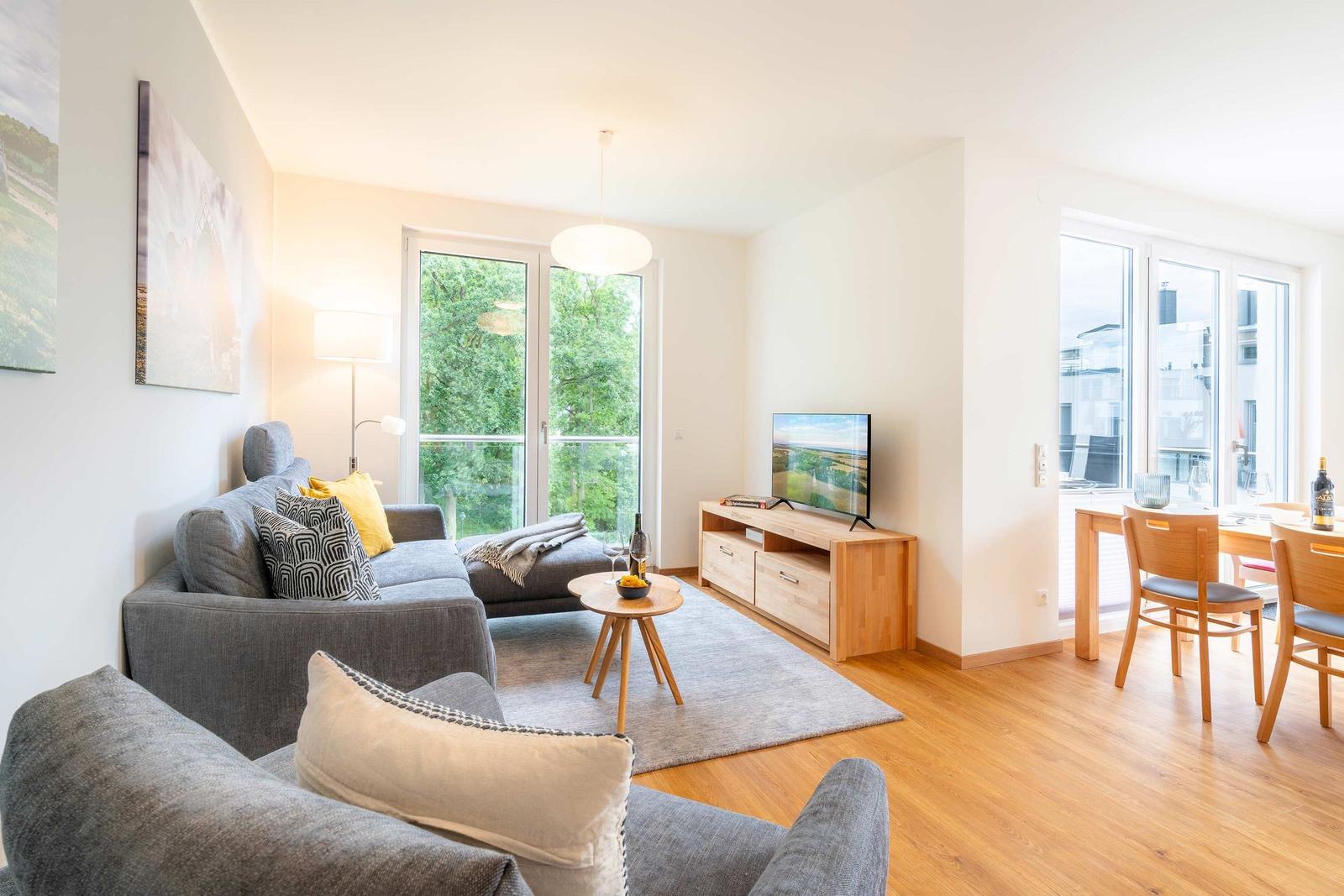 Wohnzimmer mit Sofa, Fernseher und Essbereich. Holzboden und große Fenster.