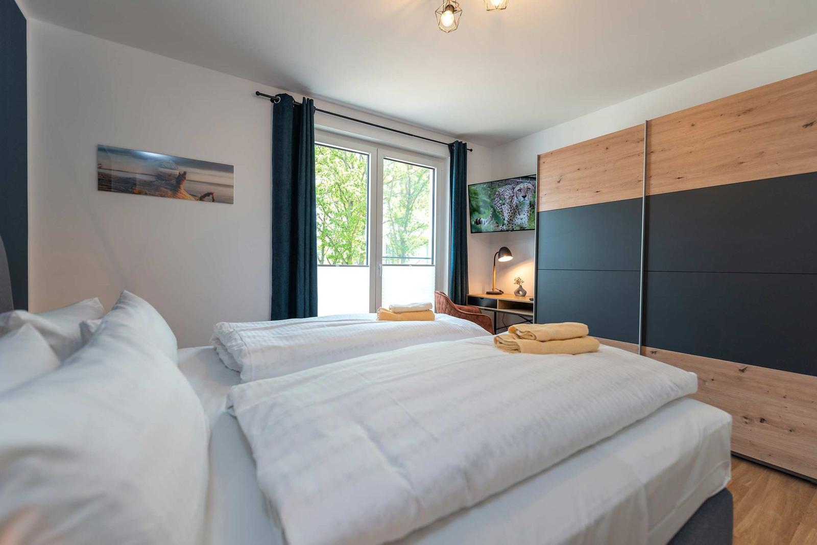 Zwei Betten mit weißen Bettwäsche, Fenster mit Blick auf Bäume und Schrank mit Holz- und schwarzen Paneelen.