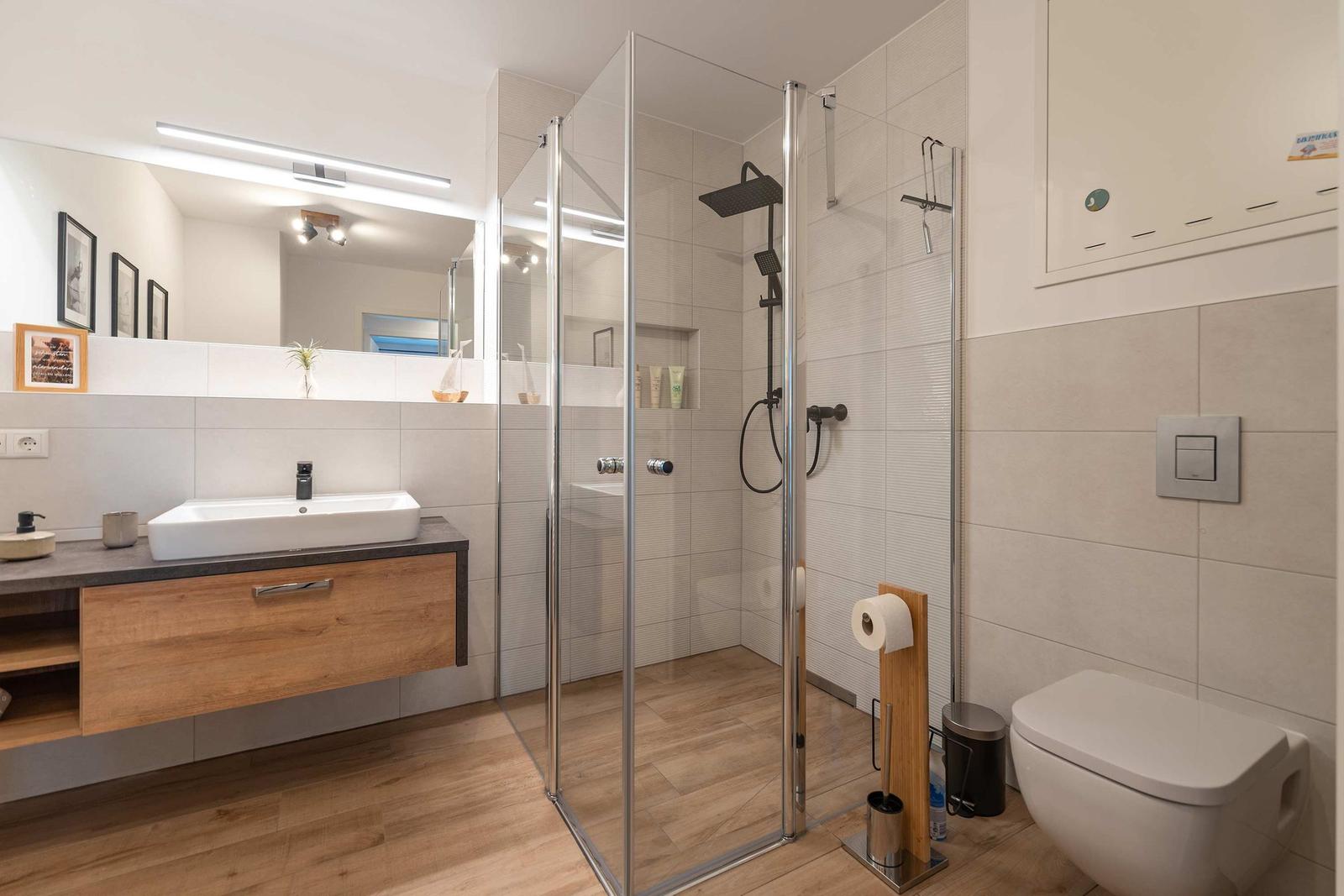 Badezimmer mit Dusche, Waschbecken und Toilette. Holzboden und hell gefliest.