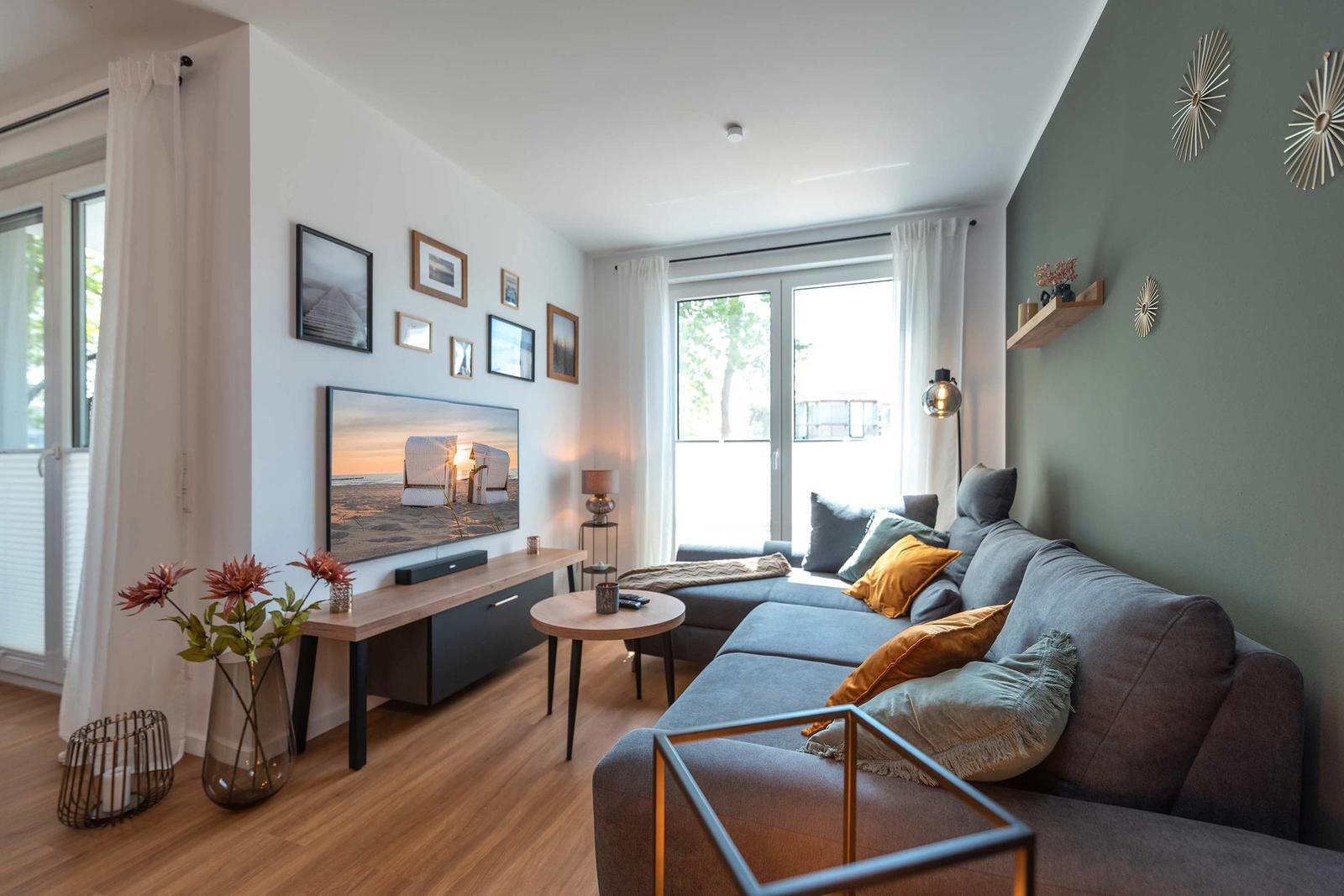 Ein Wohnzimmer mit grauem Sofa, Fernseher und großen Fenstern.
