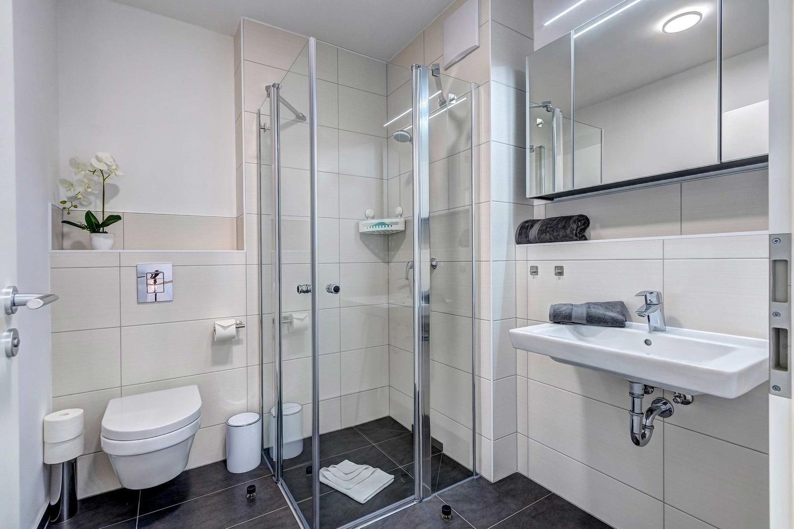 Badezimmer mit Dusche, Waschbecken und Toilette. Weiße Fliesen und dunkler Boden.