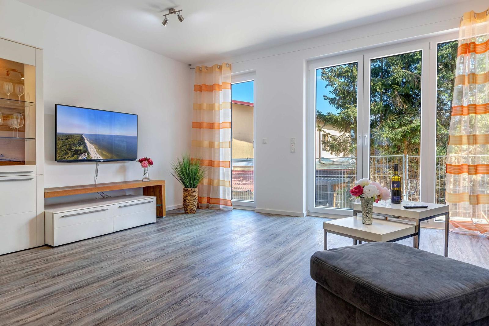 Wohnzimmer mit TV, Balkon und Holzboden. Fenster und Vorhänge sichtbar.