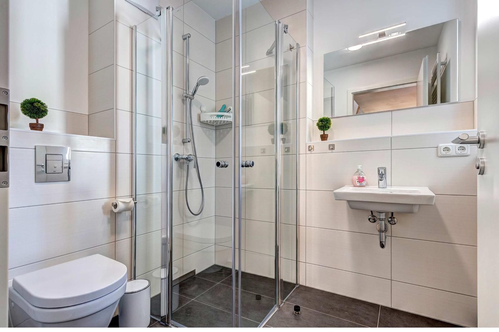 Badezimmer mit Dusche, Waschbecken und Toilette. Weiße Fliesen und Glaswand.