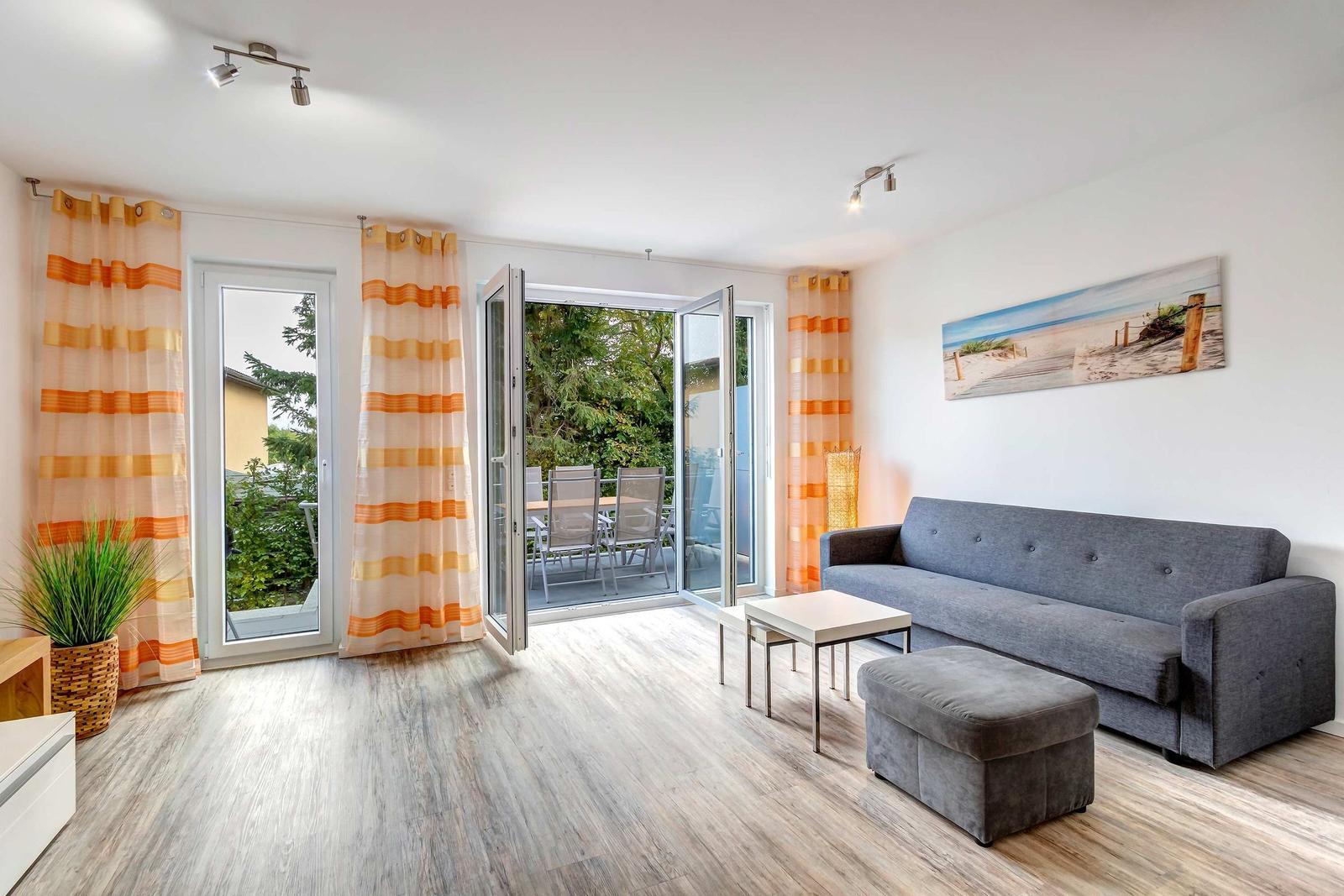 Wohnzimmer mit grauem Sofa, Tisch und Fußhocker. Durchgehende Glastüren führen auf eine Terrasse.