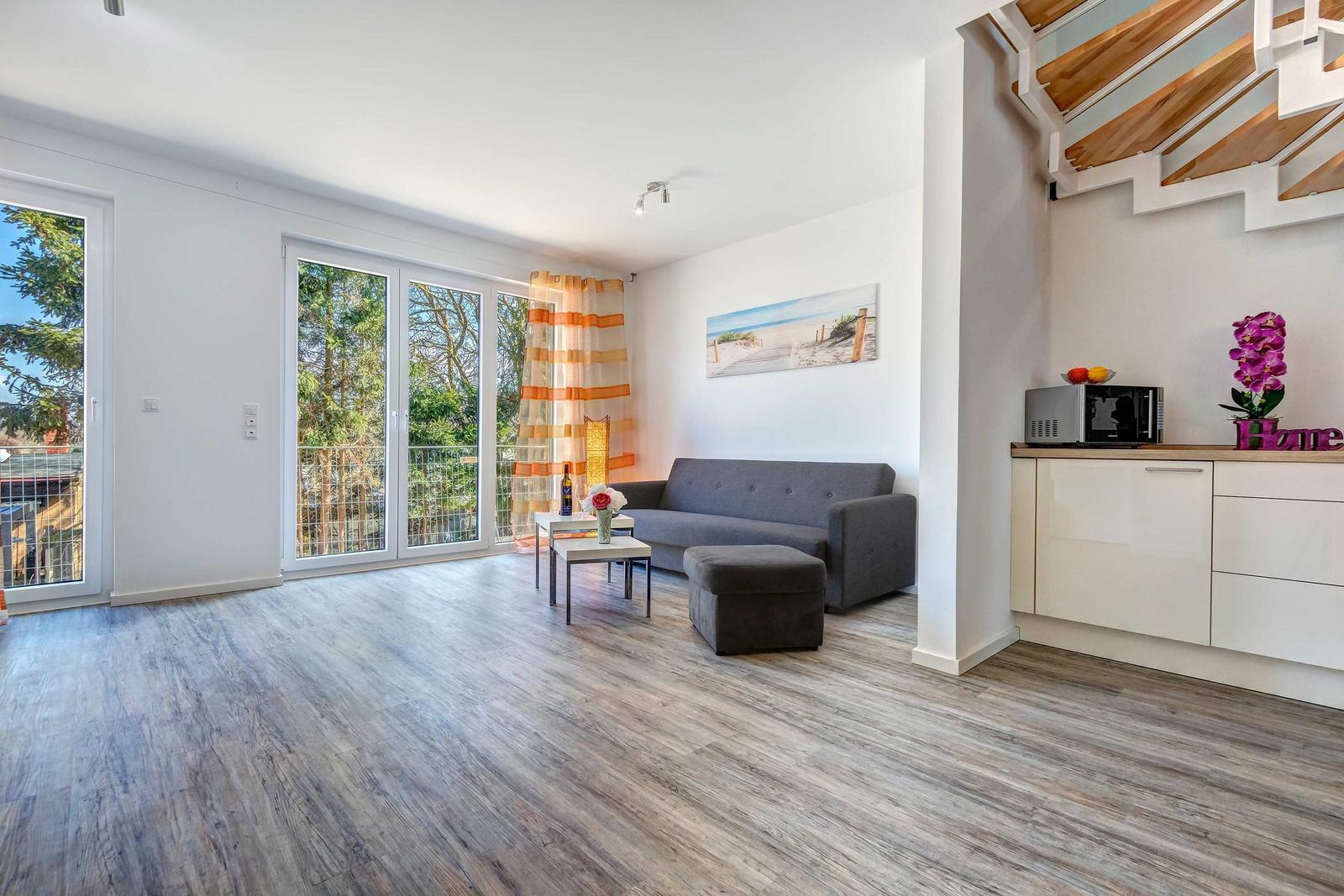 Wohnzimmer mit Sofa, Tisch und Balkontür. Küche mit Mikrowelle und Blume.