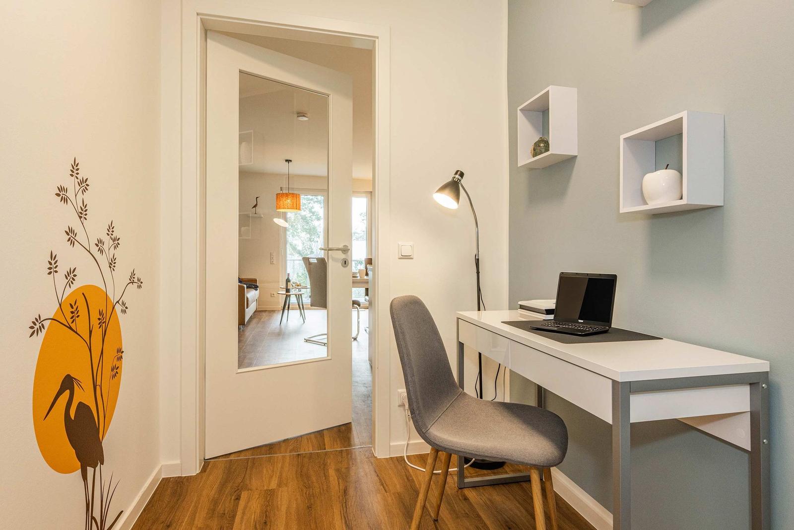 Workspace with desk, chair, and laptop. Wall decor and view into living area.