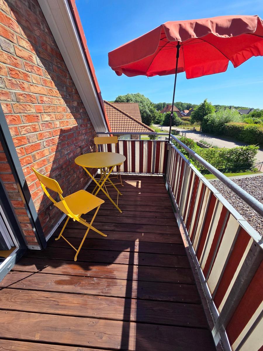 Terrasse mit gelben Stühlen, Tisch und rotem Sonnenschirm. Holzboden und Sicht auf Grünflächen.
