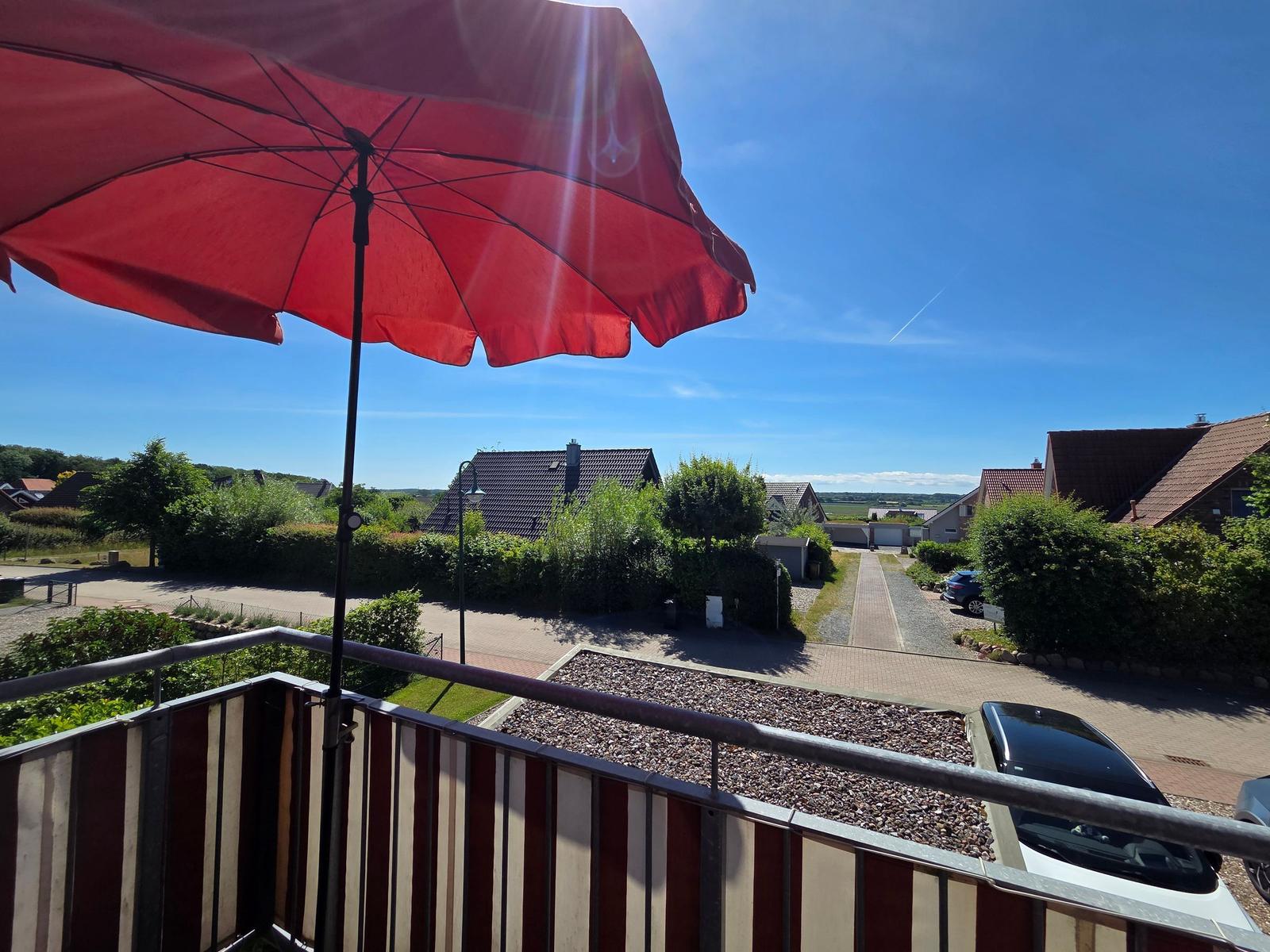 Roter Sonnenschirm auf Balkon mit Blick auf Wohnhäuser und grüne Landschaft.