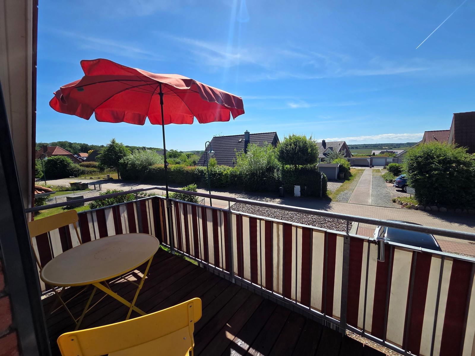 Terrasse mit rotem Sonnenschirm und gelben Stühlen. Blick auf Wohngebiet und grüne Landschaft.