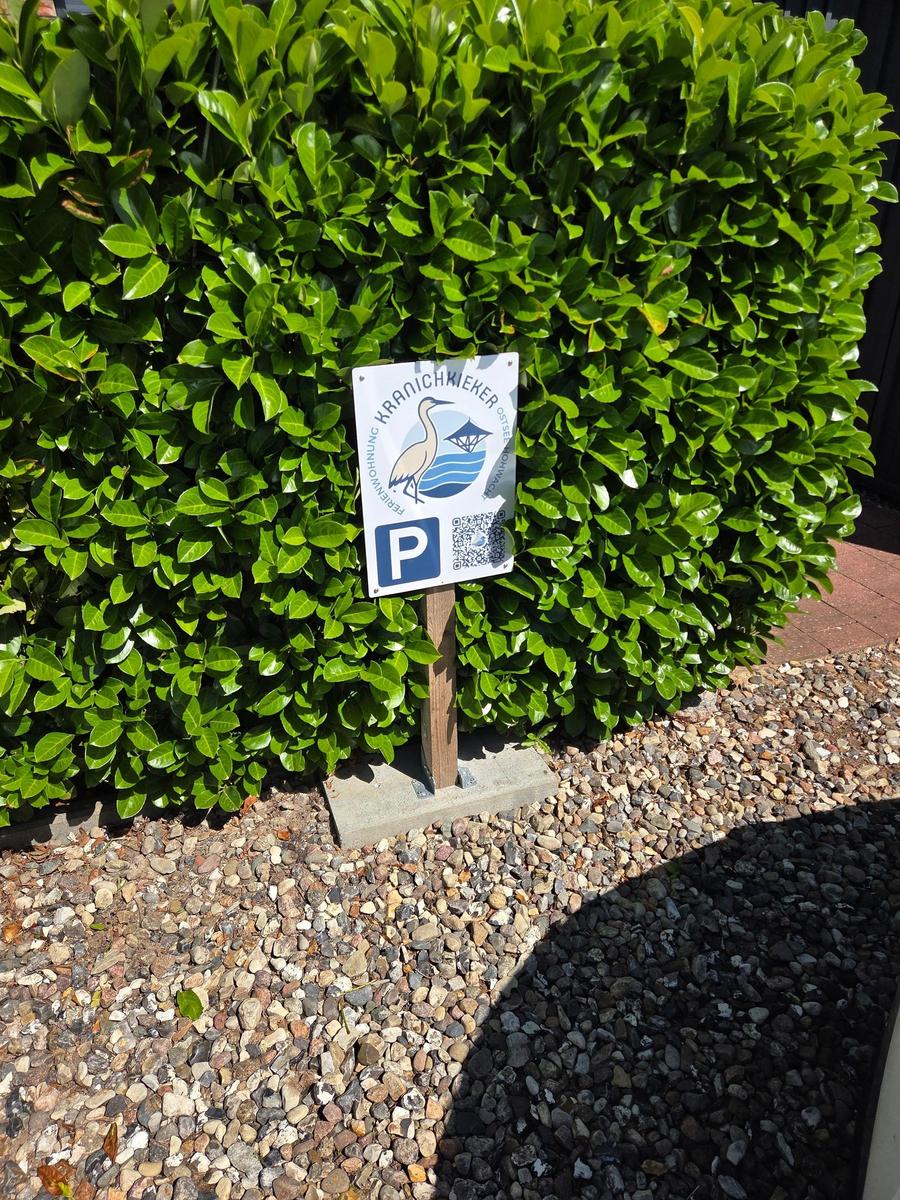 Parkplatzschild vor grüner Hecke mit QR-Code und Logo