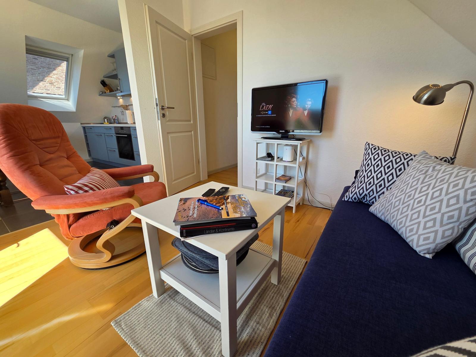 Wohnbereich mit Sofa, Fernseher und Sessel. Küche im Hintergrund sichtbar.