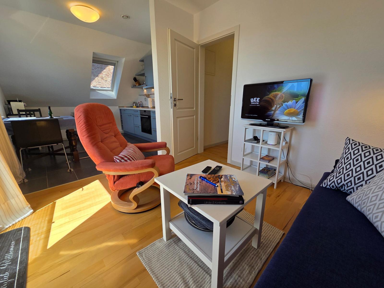 Wohnzimmer mit Sofa, Fernseher und orangefarbenem Sessel. Küche und Schlafzimmer sind sichtbar.
