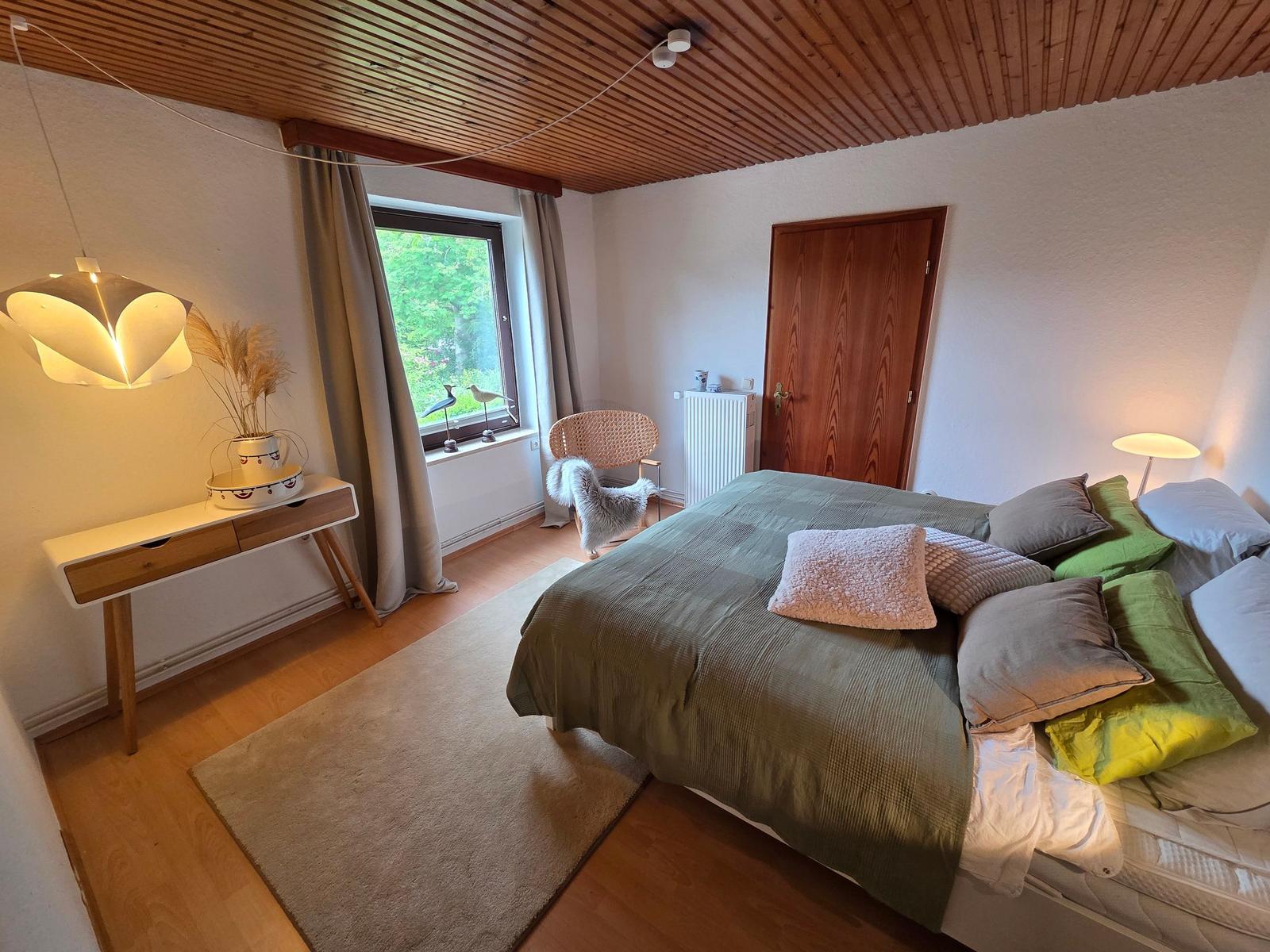 Schlafzimmer mit Bett, Fenster, Schreibtisch und Holzdecke.